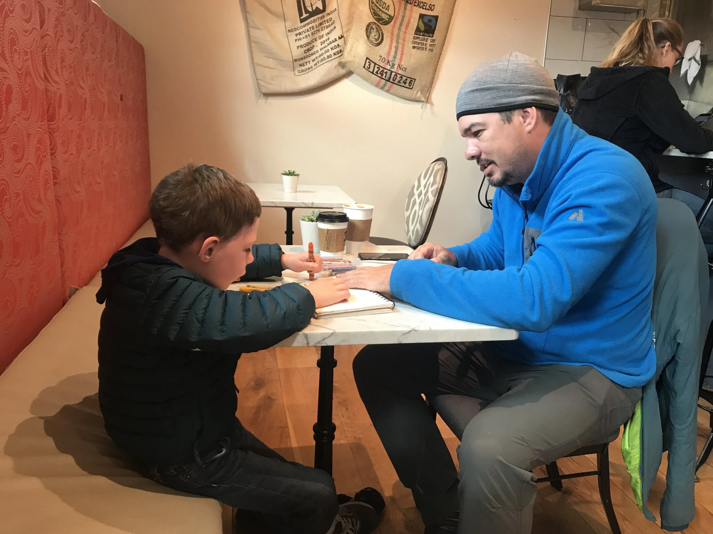 A boy working on schoolwork at a table while his dad  in a blue fleece helps alongside him.