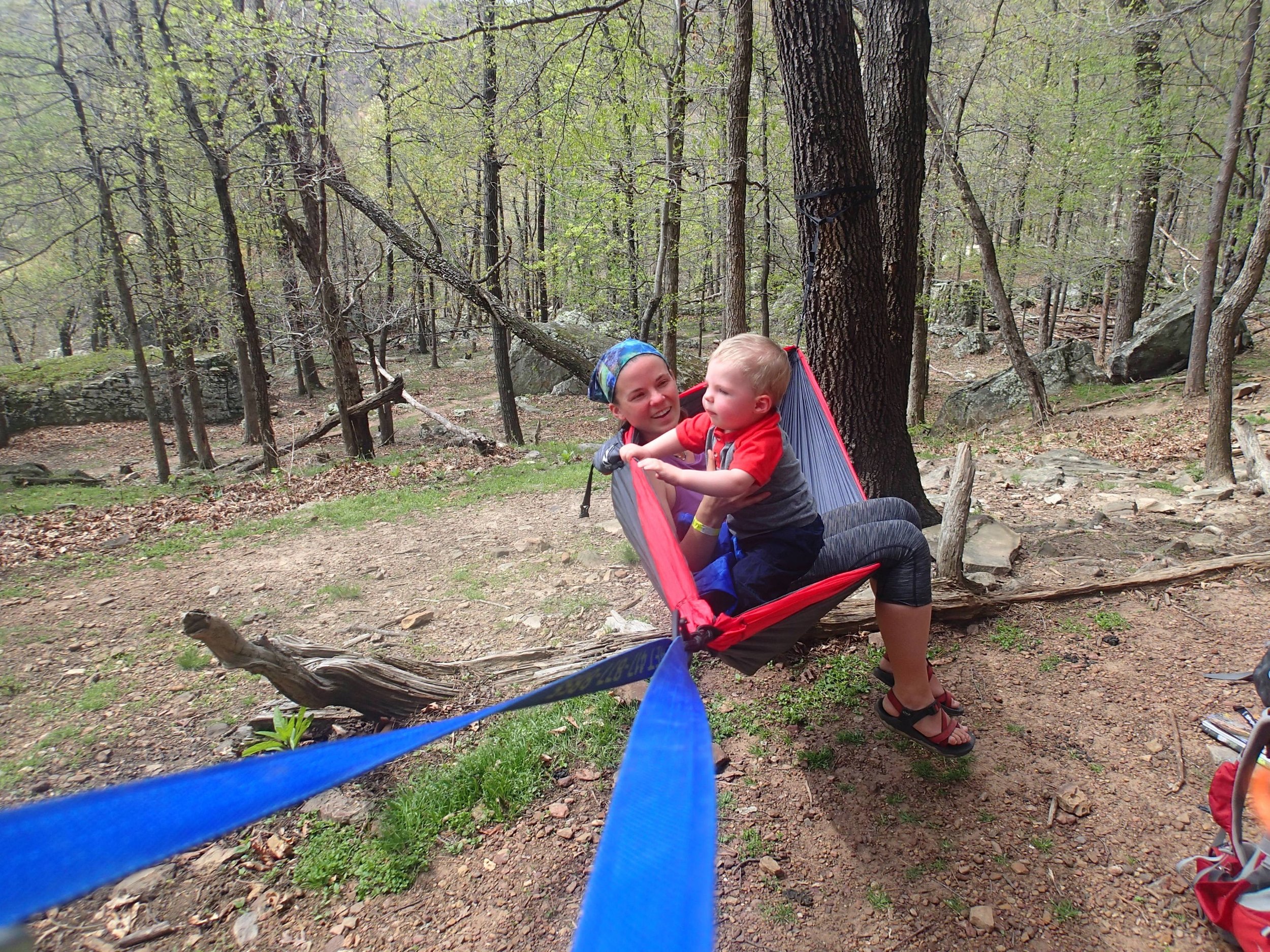 Mom and a child sit together in a camping hammock strung between trees in the woods.