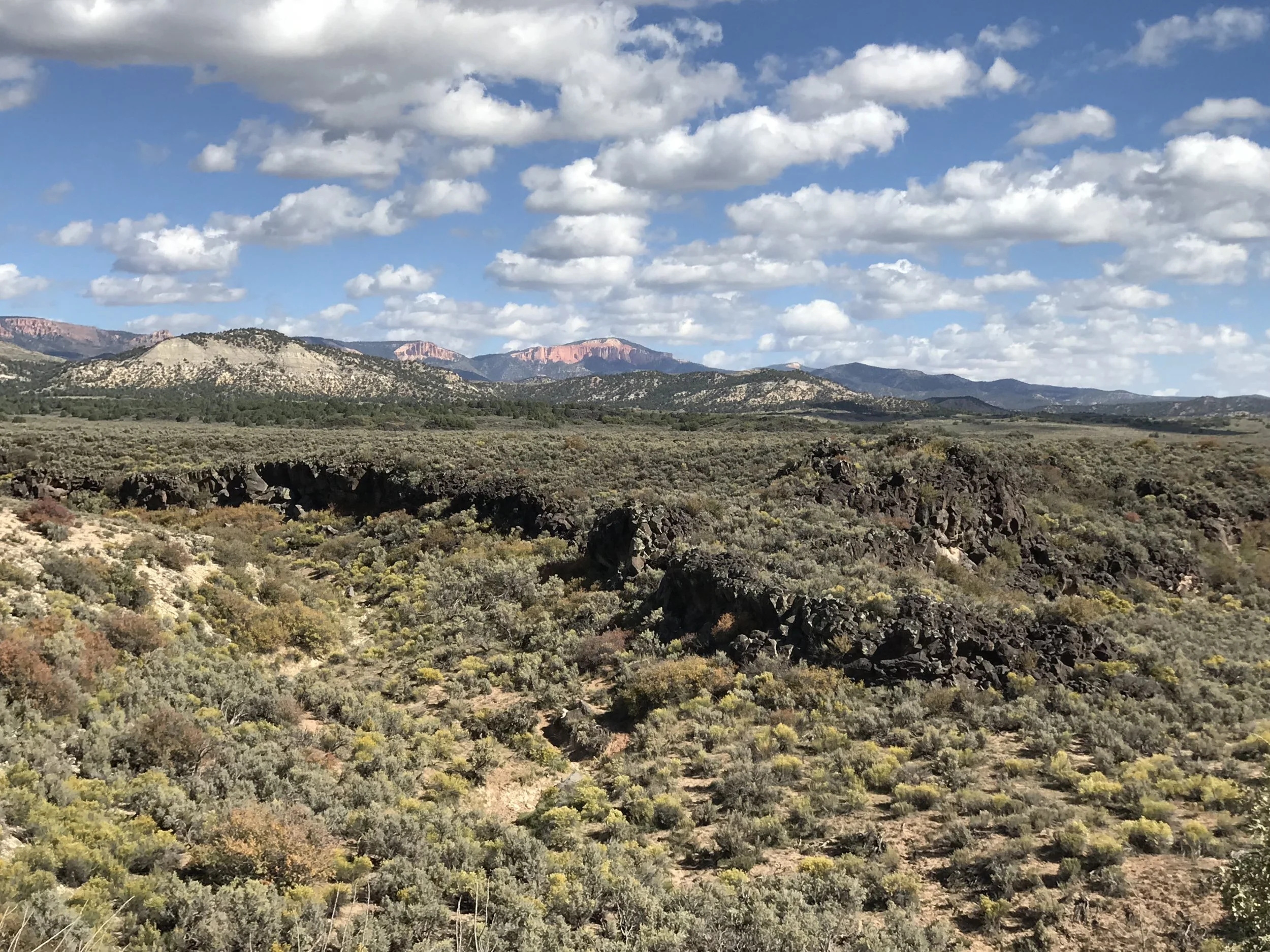 A wide sagebrush valley in southern Utah with pale and pink sandstone cliffs on the horizon under tall clouds.
