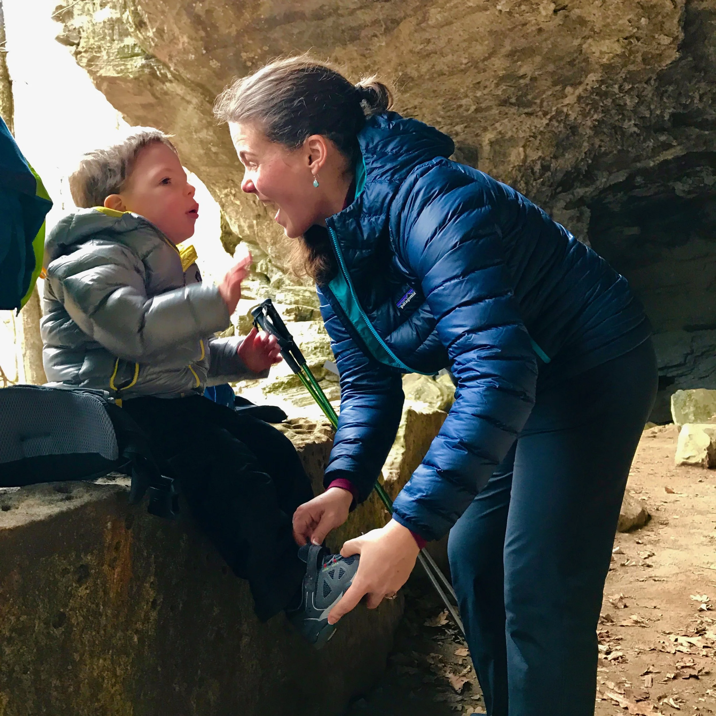 A mom helps her young son adjust his shoes for better comfort as he sits on a rock, eating a snack, during a hike.