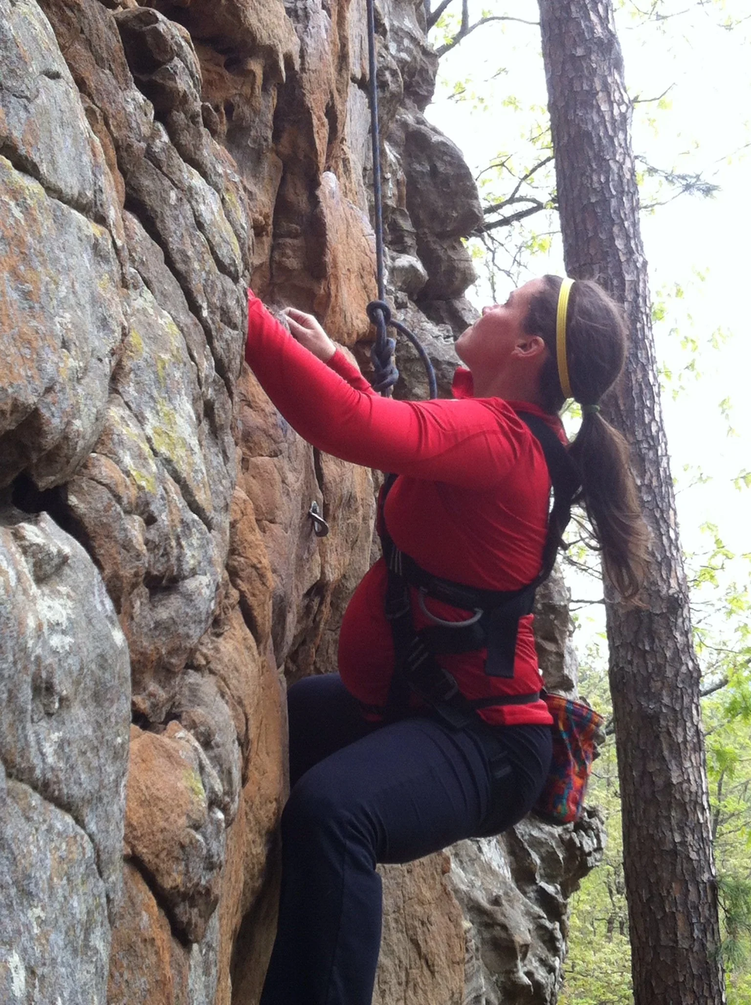 A pregnant climber in a red top on a rock face wearing a maternity climbing harness.