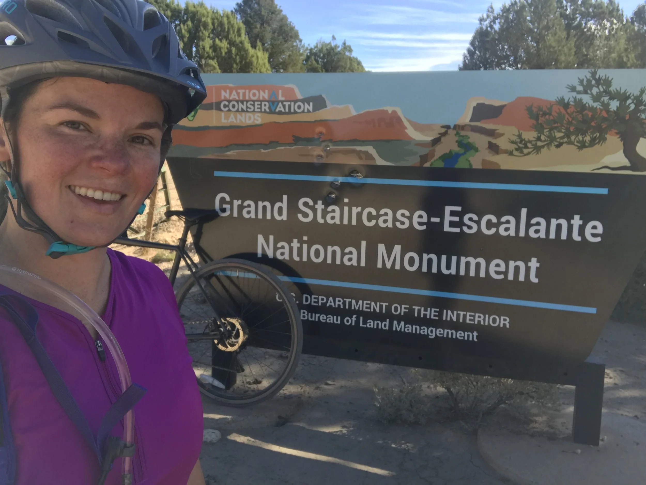 A smiling cyclist beside the Grand Staircase-Escalante National Monument entrance sign.