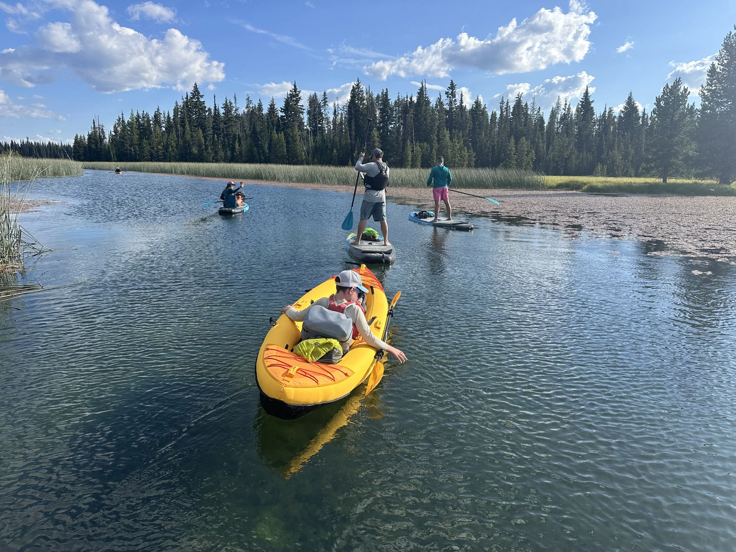 A child sitting in a yellow inflatable kayak is towed along on a calm, clear mountain lake by paddleboarders.