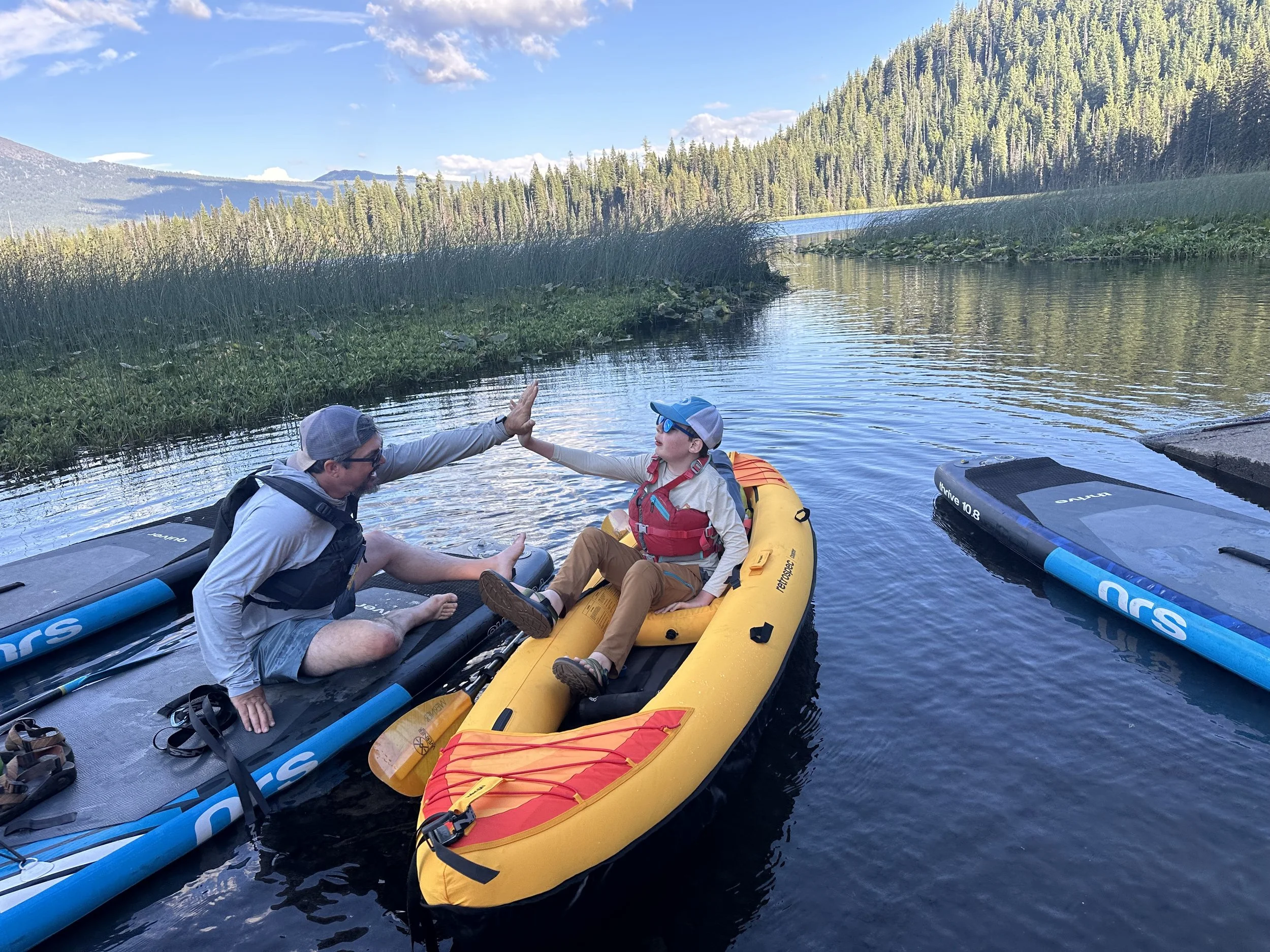 An adult and a child high-fiving from a yellow inflatable kayak on a calm river, paddleboards alongside.