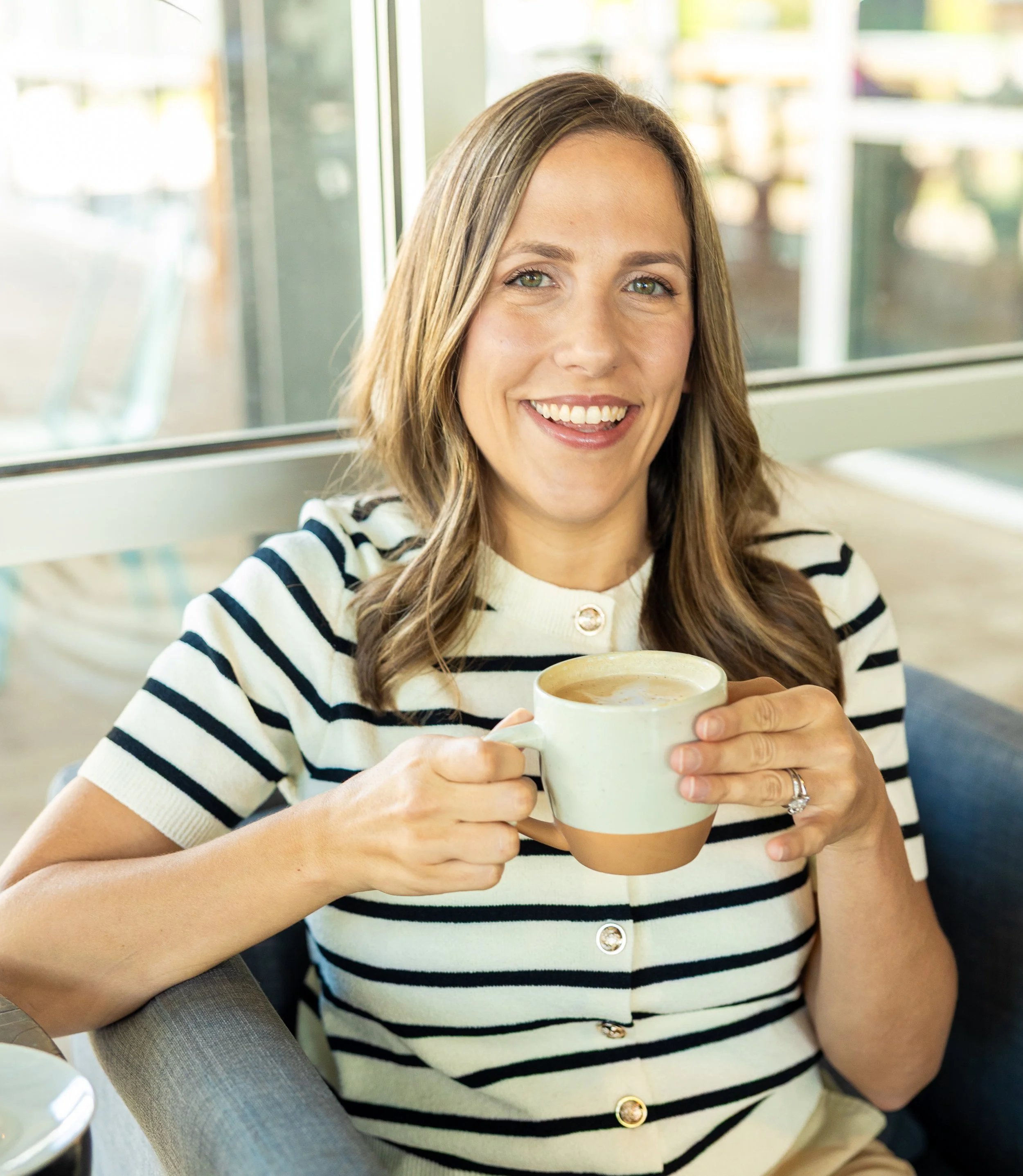 A woman with long light brown hair and green eyes holding a beige mug with a hot beverage inside, smiling in a casual setting with large windows and greenery outside.