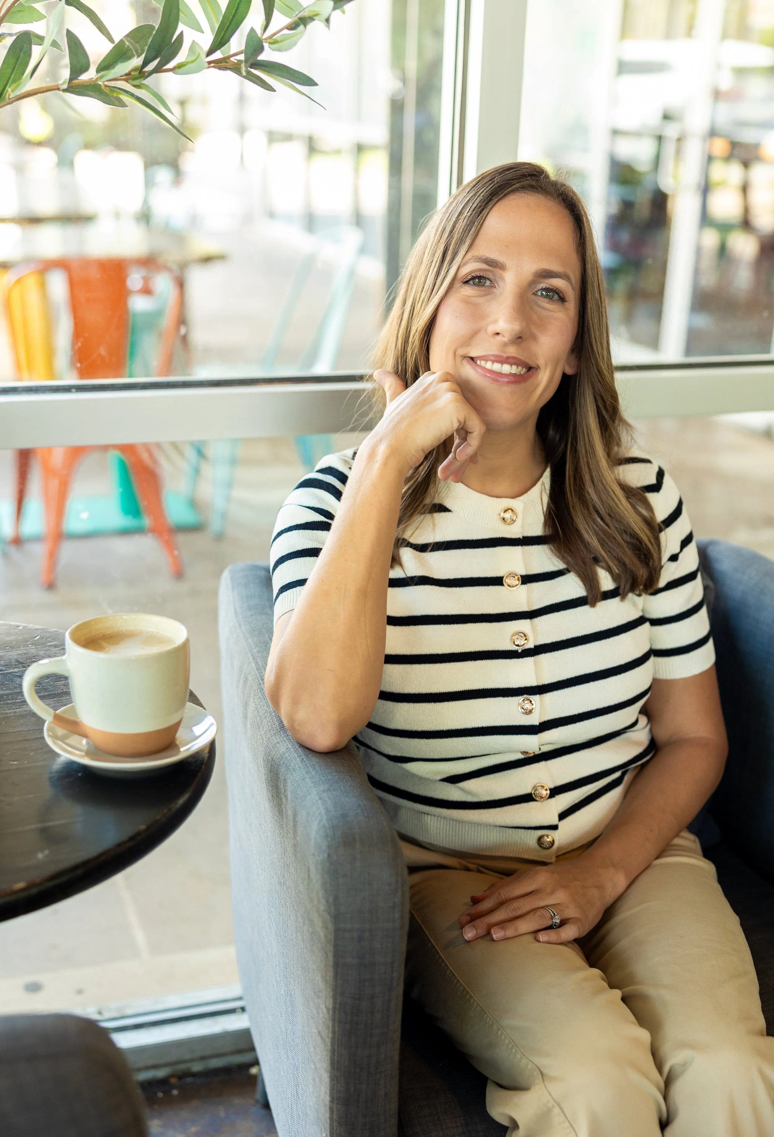 A woman sitting in a coffee shop with a cup of coffee on the table. She has long brown hair, is smiling, and is wearing a striped shirt and khaki pants. The background is bright with large windows and colorful chairs outside.