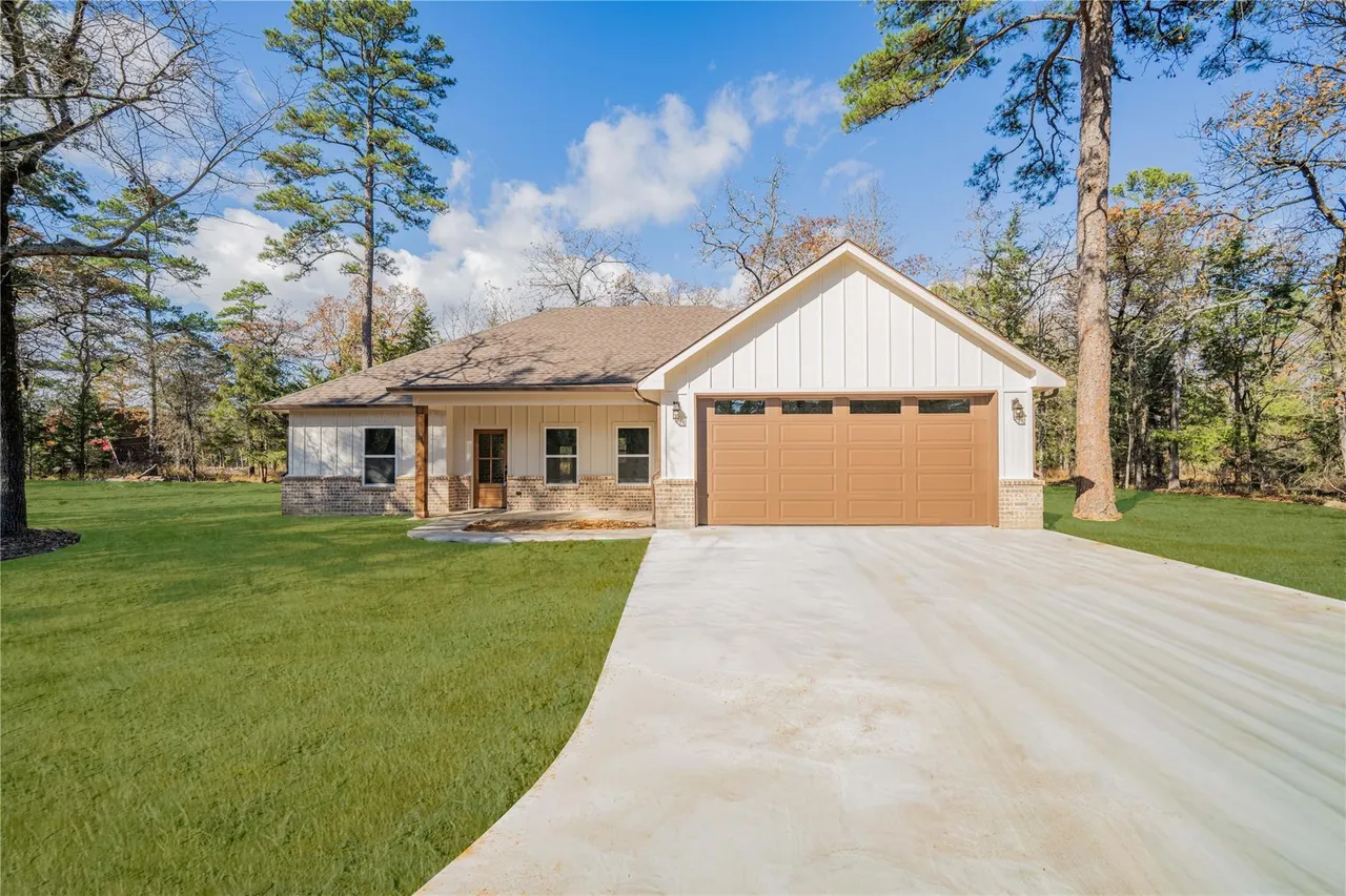 A new house with a brown garage door, white siding, and a small front porch, surrounded by a well-maintained green lawn and tall trees, under a partly cloudy blue sky.