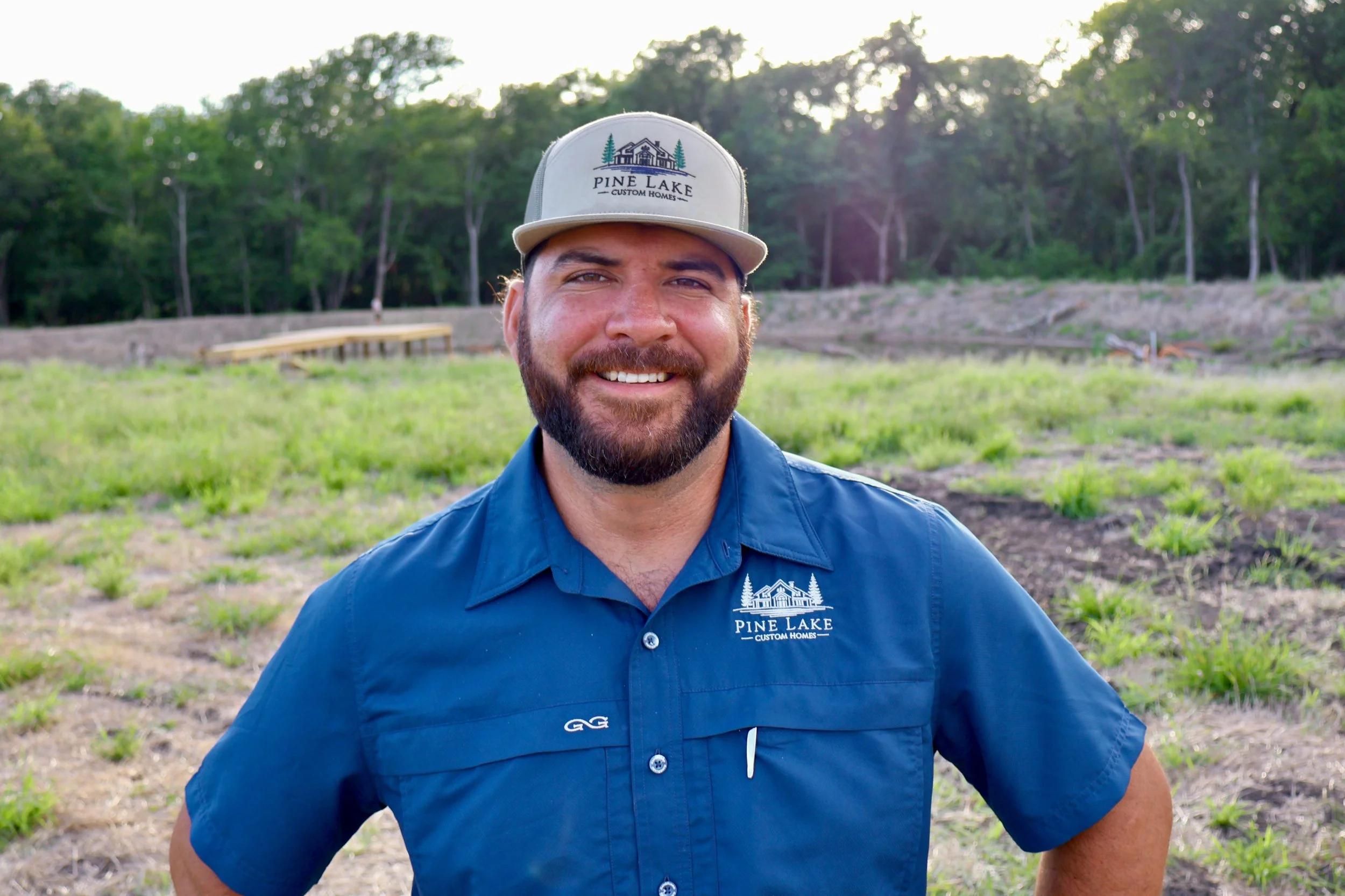 Trevor Sandoval wearing a blue shirt and a gray cap with the logo 'Pine Lake Custom Homes' on it, with a background of green trees and a partly cloudy sky.