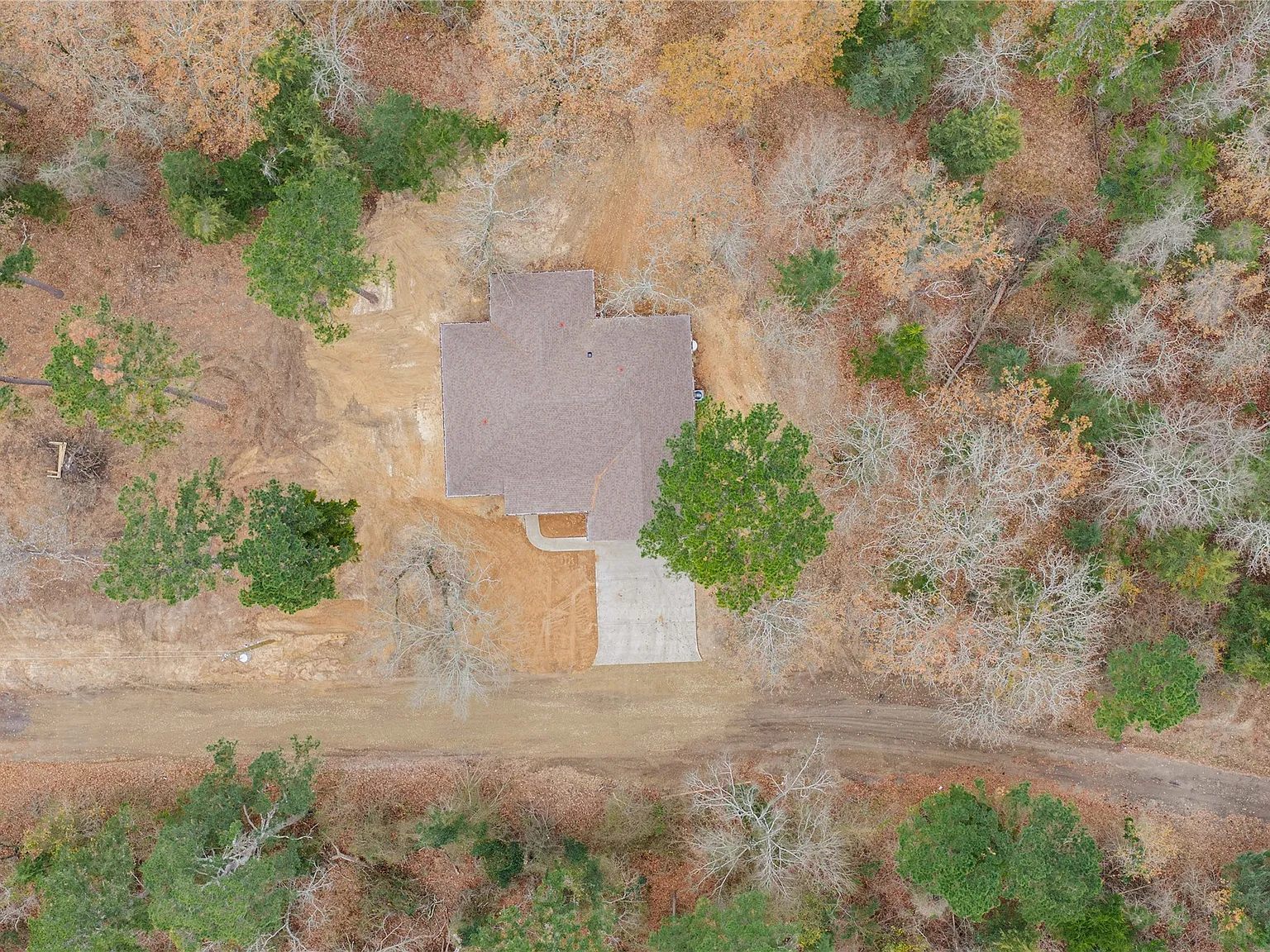 An aerial top-down view of a house with a gray roof and a driveway, surrounded by trees with autumn-colored leaves, some of which are bare.