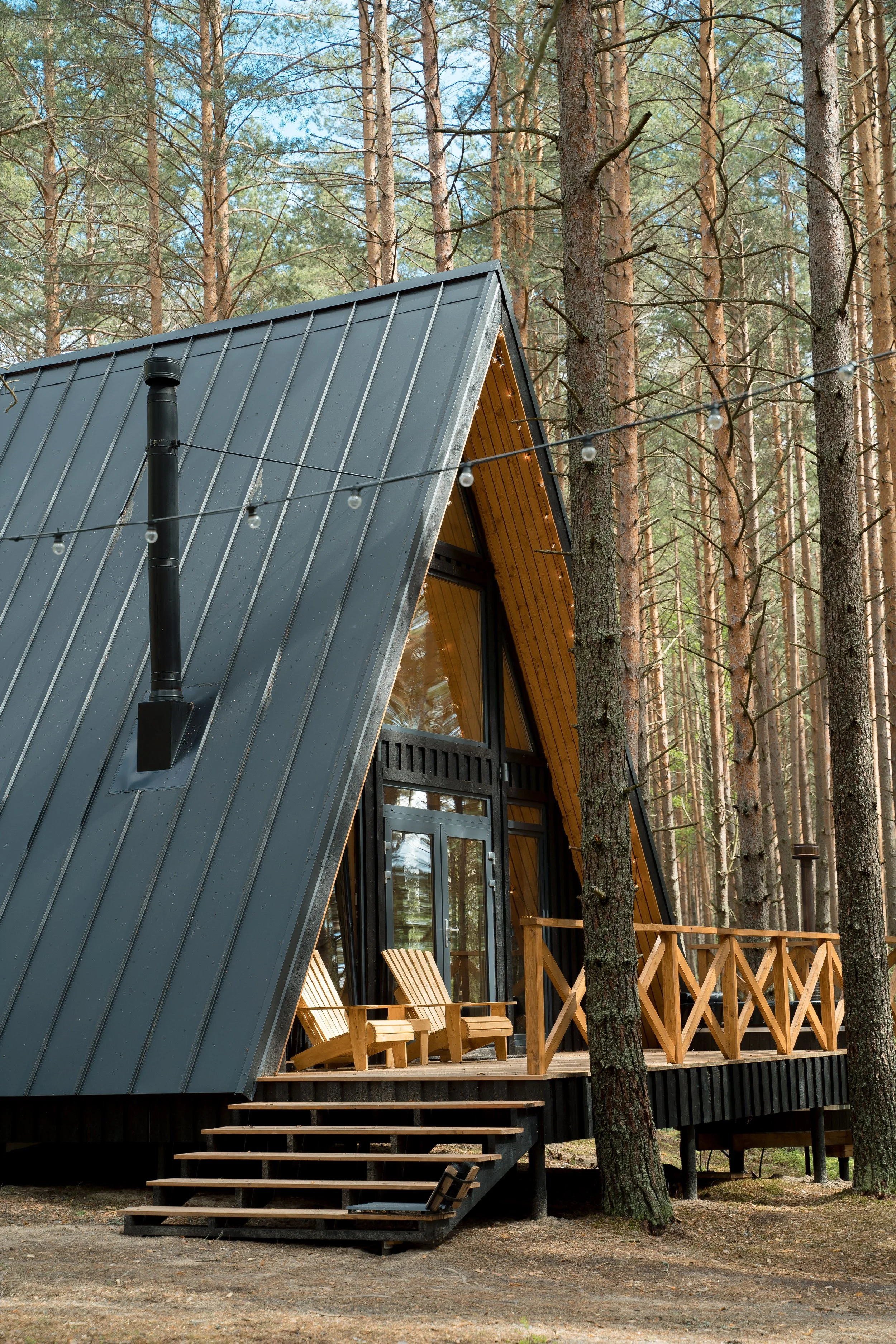 A modern A-frame cabin with a dark metal roof, situated in a wooded forest with tall pine trees. The cabin has a wooden balcony with chairs and string lights hanging above, and a view of the trees surrounds the area.