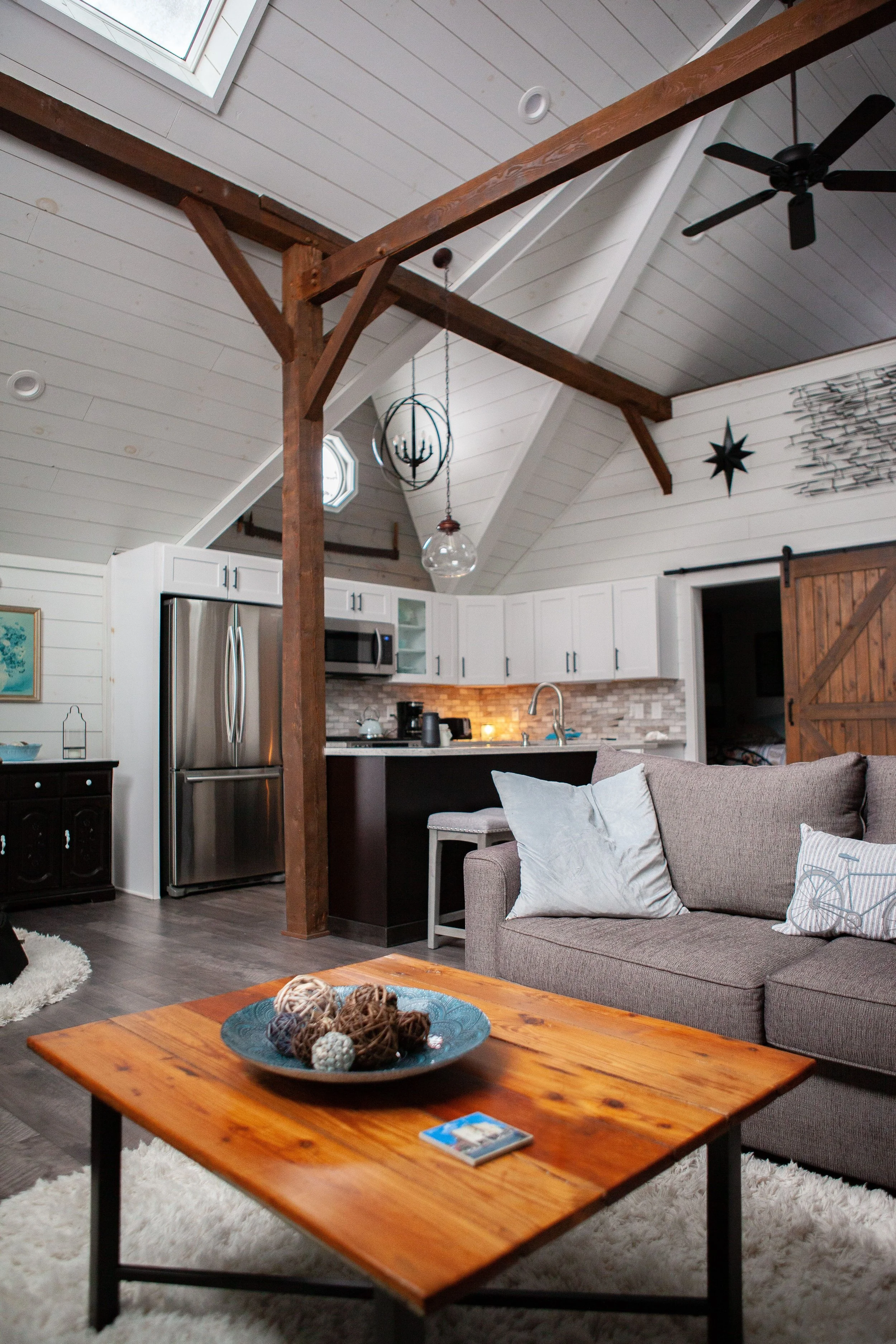Living room with a gray sofa, white pillows, a wooden coffee table with a decorative bowl of balls, a white rug, and an open kitchen area with white cabinets and stainless steel appliances. The ceiling has wooden beams, a ceiling fan, and a hanging chandelier, with small octagonal windows and a wall decoration.