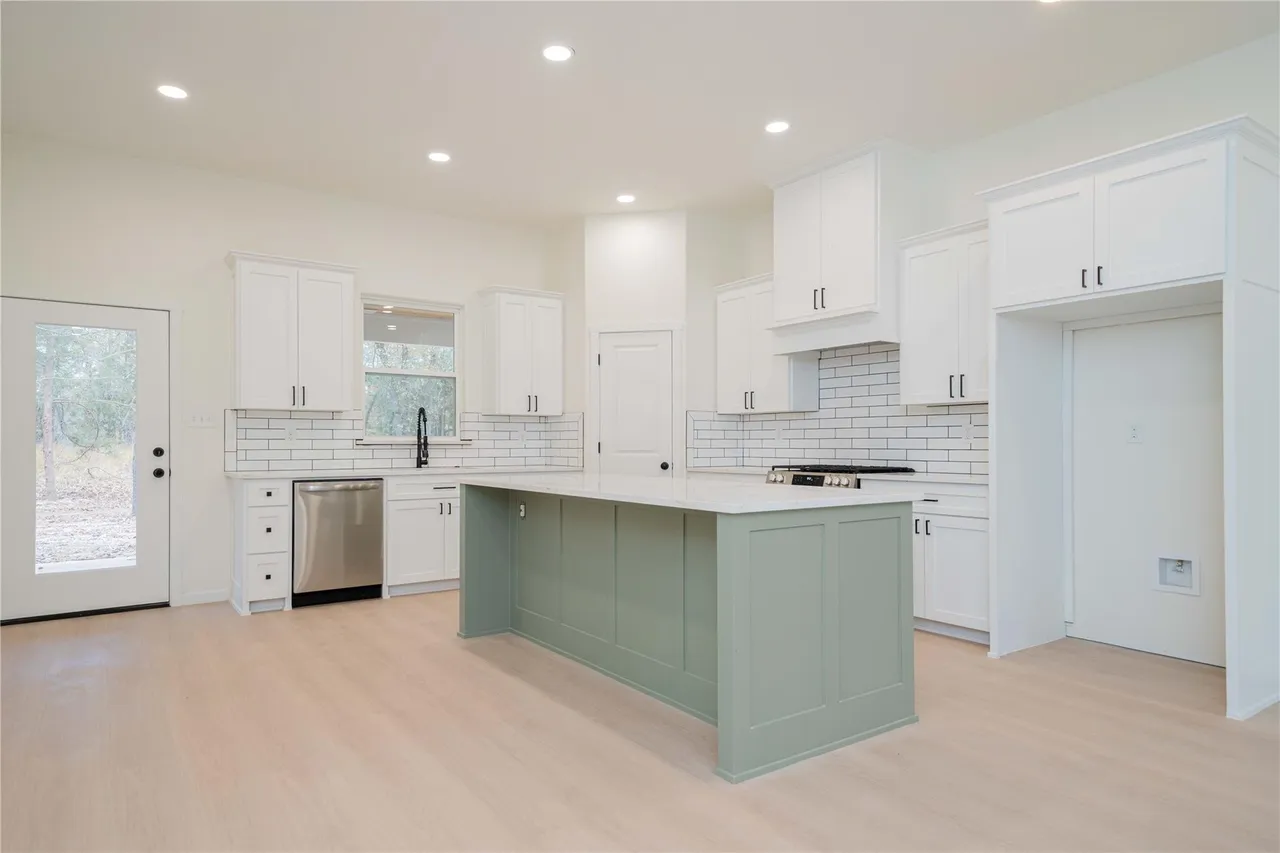 Modern kitchen with white cabinets, a green island, white subway tile backsplash, stainless steel dishwasher, and black hardware, with a door to the outside.