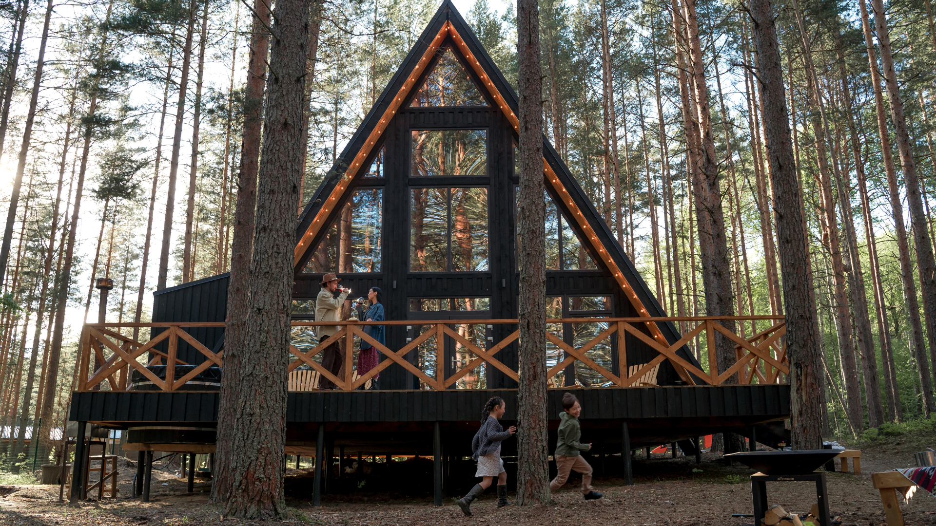 A black A-frame cabin with large windows and string lights, built among tall pine trees. People are standing on the balcony, with children walking in front. The scene is set in a forest during daytime.