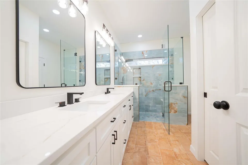 Modern bathroom with double vanity, black faucets, large mirrors, and a glass-walled shower with blue and brown tile.