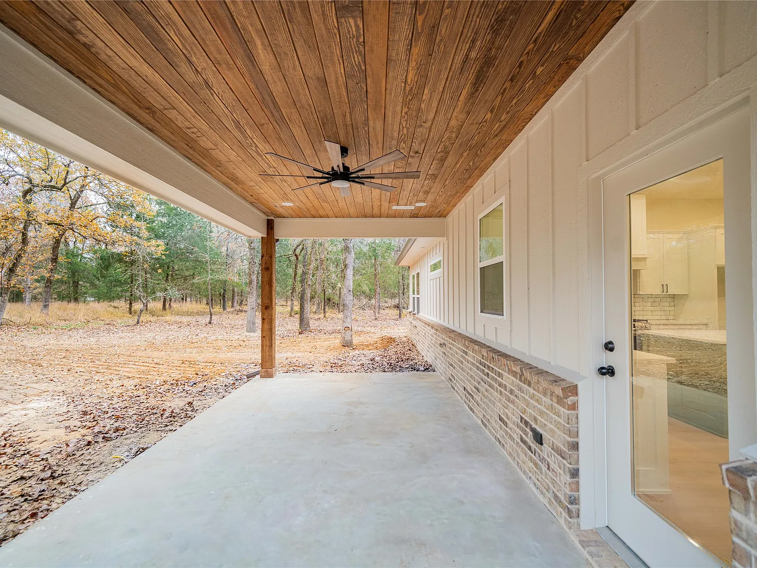 Covered patio area with concrete floor, wooden ceiling, ceiling fan, and door leading to the interior of a house, with a view of a wooded yard.