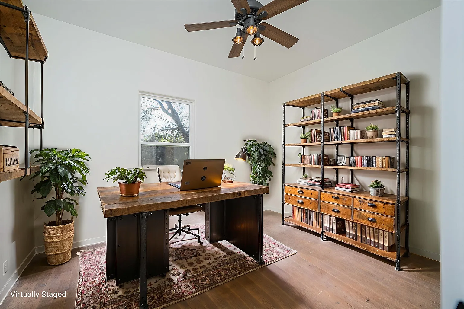 Home office with a wooden desk, a white chair, a laptop, a large bookshelf with books and plants, and potted plants near the window, illuminated by natural light