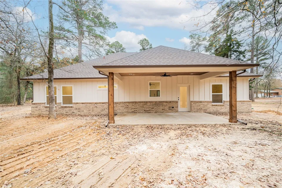 Rear view of a newly built single-story house with white exterior walls, a brick foundation, and a covered patio with wooden support beams, surrounded by a dirt yard and trees.