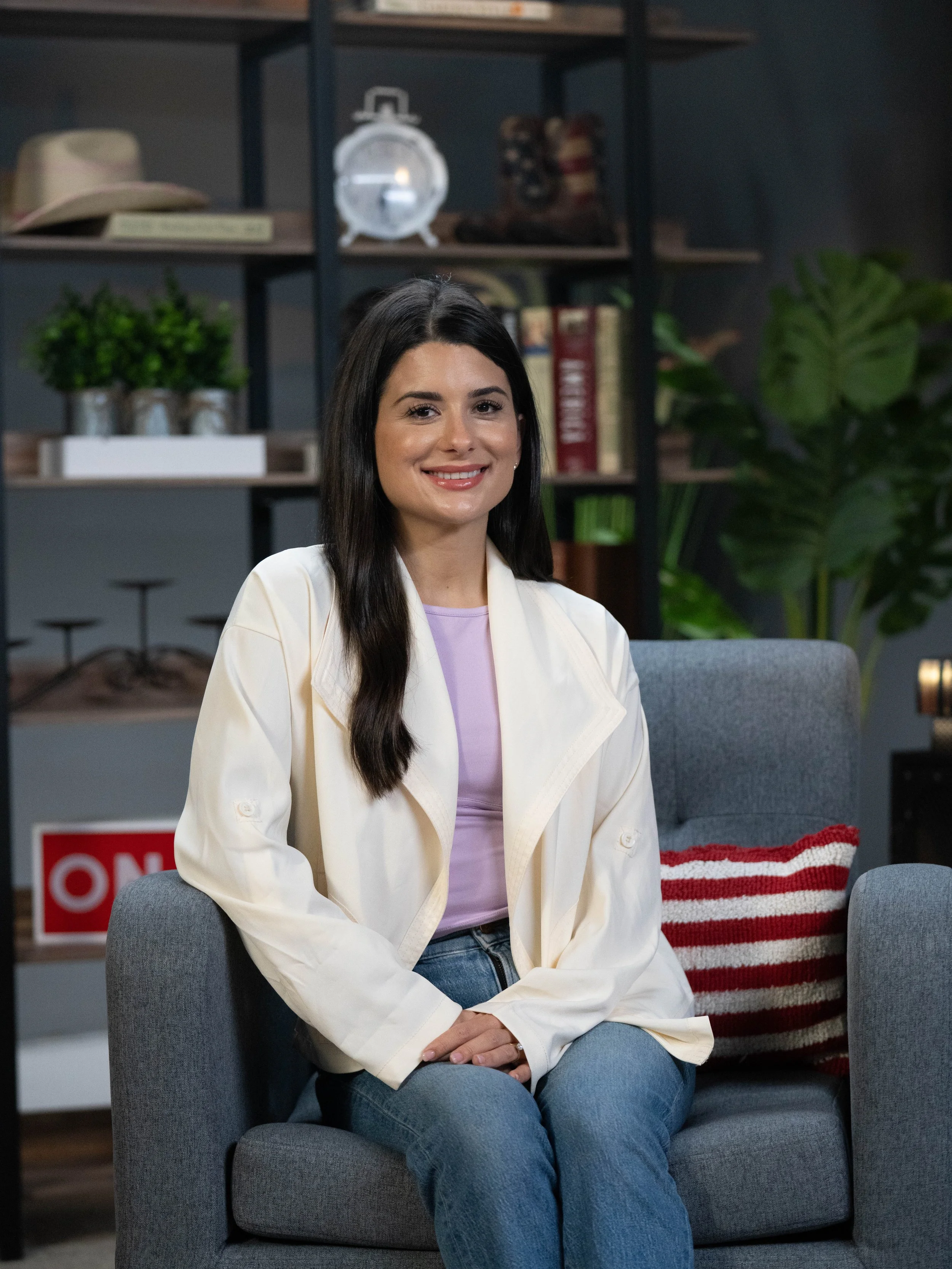 A woman with long dark hair and light skin sitting on a gray armchair in a living room. She is smiling, wearing a cream-colored jacket over a lavender top, and blue jeans. Behind her, there are shelves with books, plants, and decorative items.