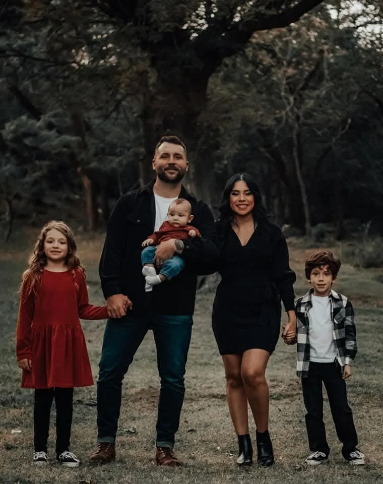 A smiling family of four with two children standing together outdoors under a tree canopy in Lubbock Texas