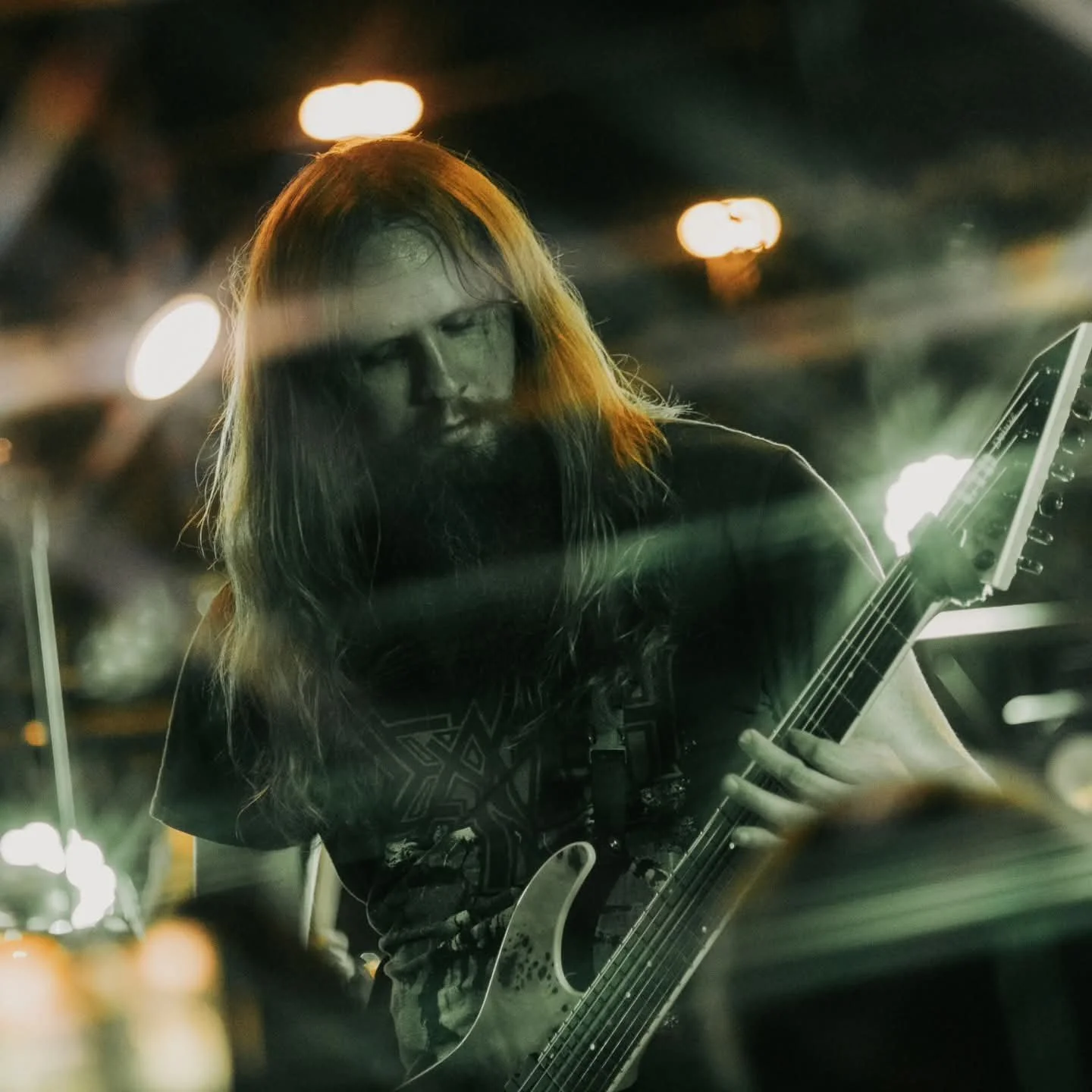 A long haired guitarist with a beard playing an electric guitar under dramatic green stage lights during a live concert in Lubbock Texas