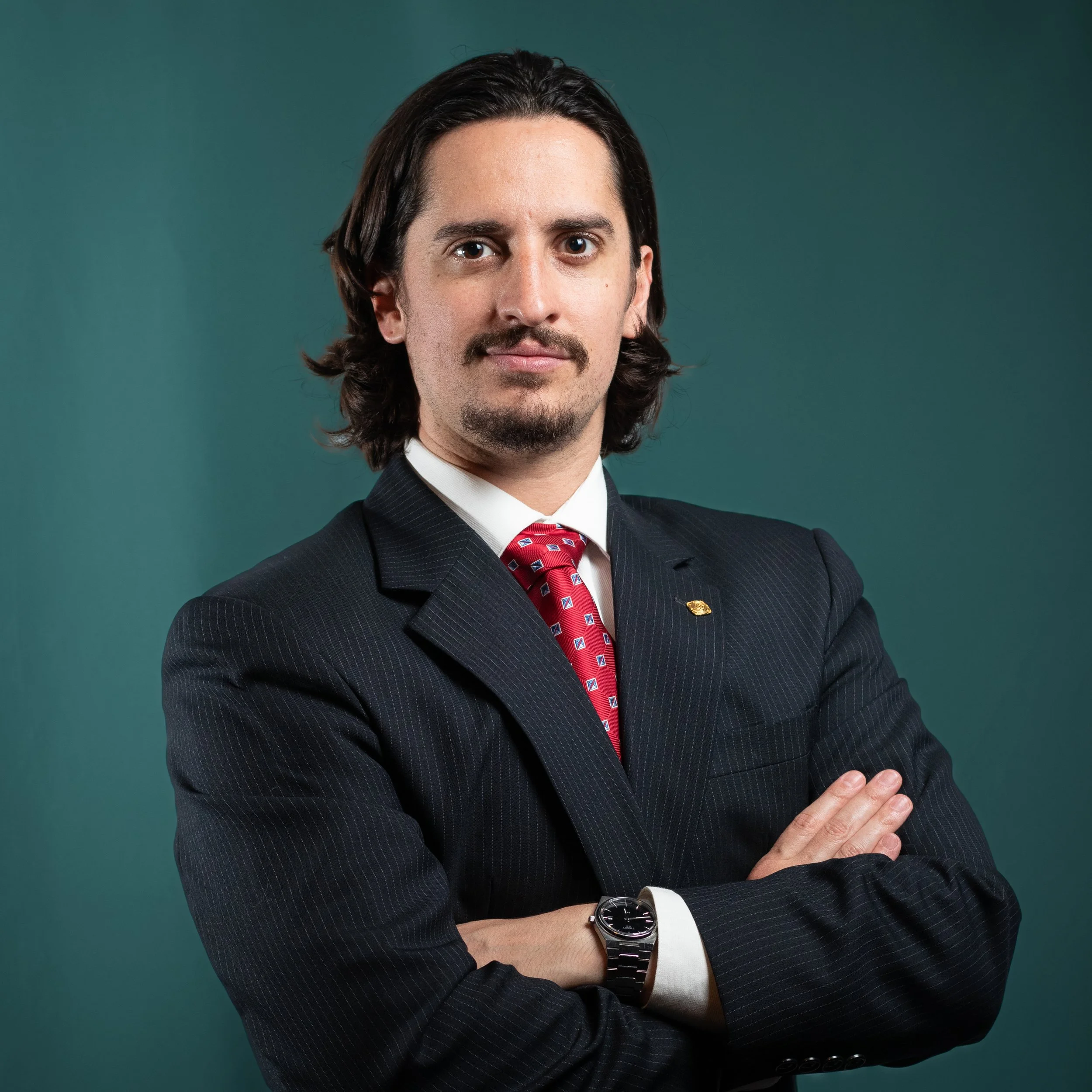A professional studio headshot of a confident man in a dark suit and red tie against a teal background in Lubbock Texas