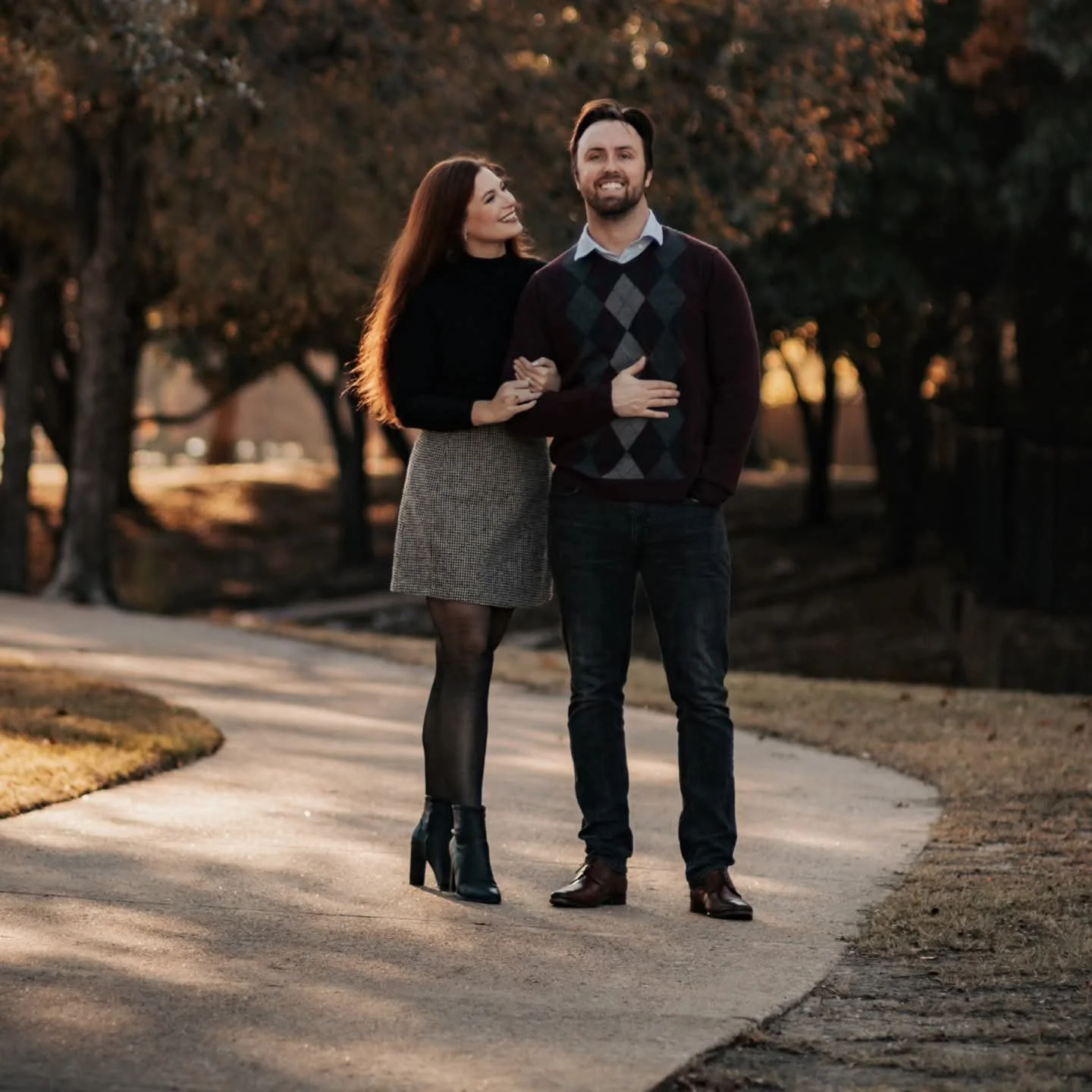 A smiling man in a patterned sweater vest and a woman in a grey skirt standing together on a paved walkway in a park surrounded by trees in Dallas Texas