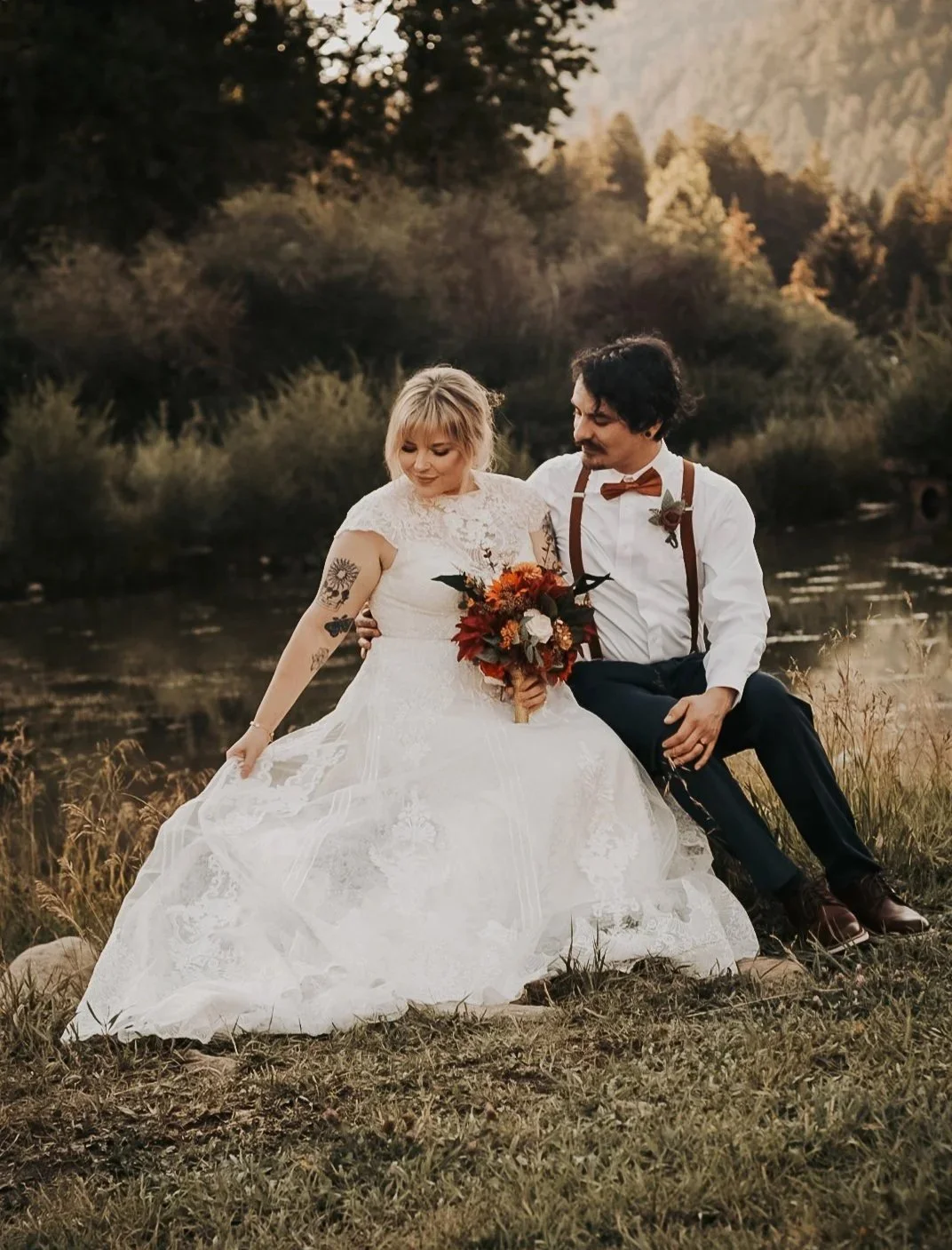 A bride in a lace wedding dress holding a vibrant bouquet and a groom in a bowtie and suspenders sitting together in a field at sunset in Lubbock Texas