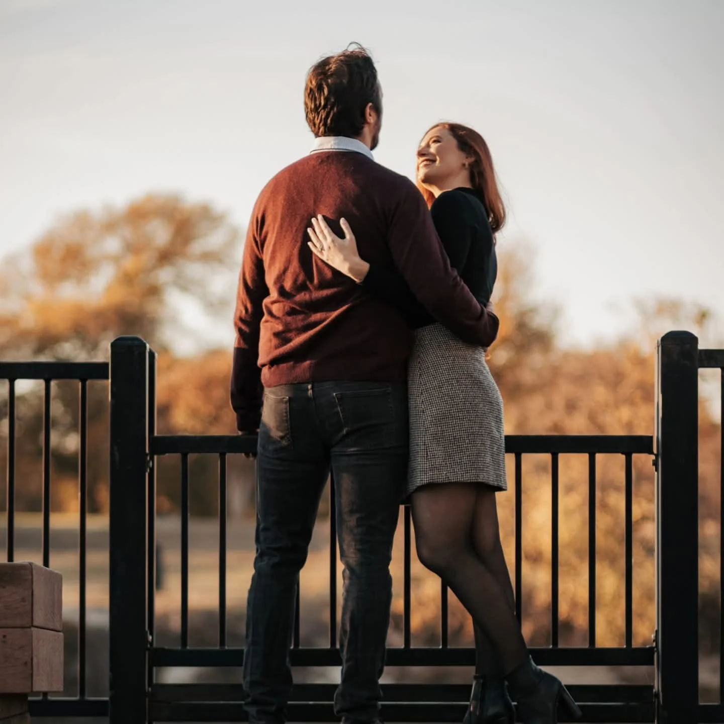 A smiling man in a dark red sweater and a woman in a grey dress embracing on a wooden bridge at sunset in Dallas Texas