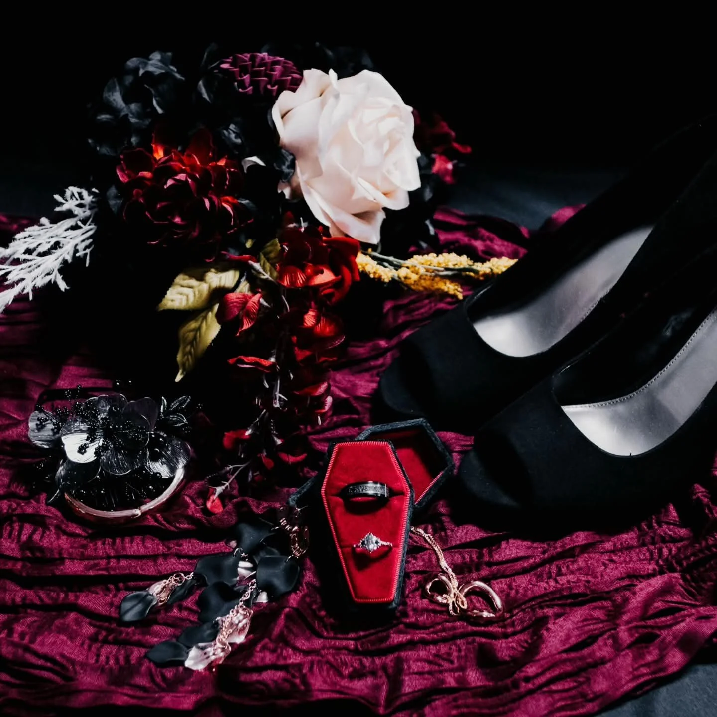 A flat lay of moody wedding details featuring black high heels, a red coffin ring box, and dark floral arrangements on a wine red velvet fabric backdrop in Lubbock Texas