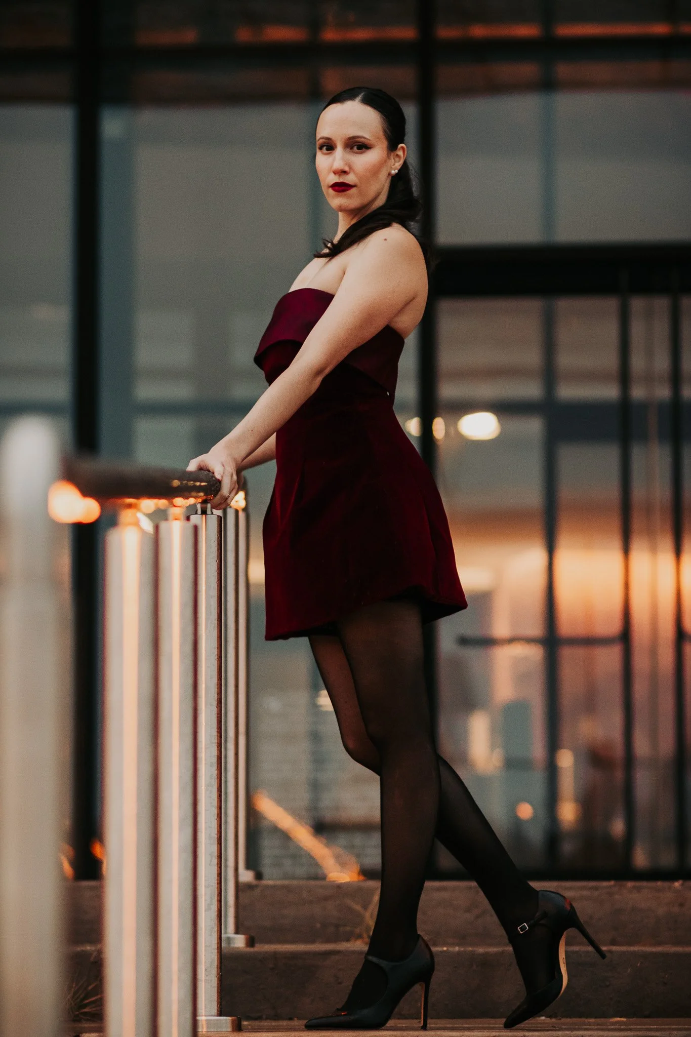 A portrait of a elegant model wearing a dark red dress and black tights leaning against a metallic handrail at dusk in Lubbock Texas