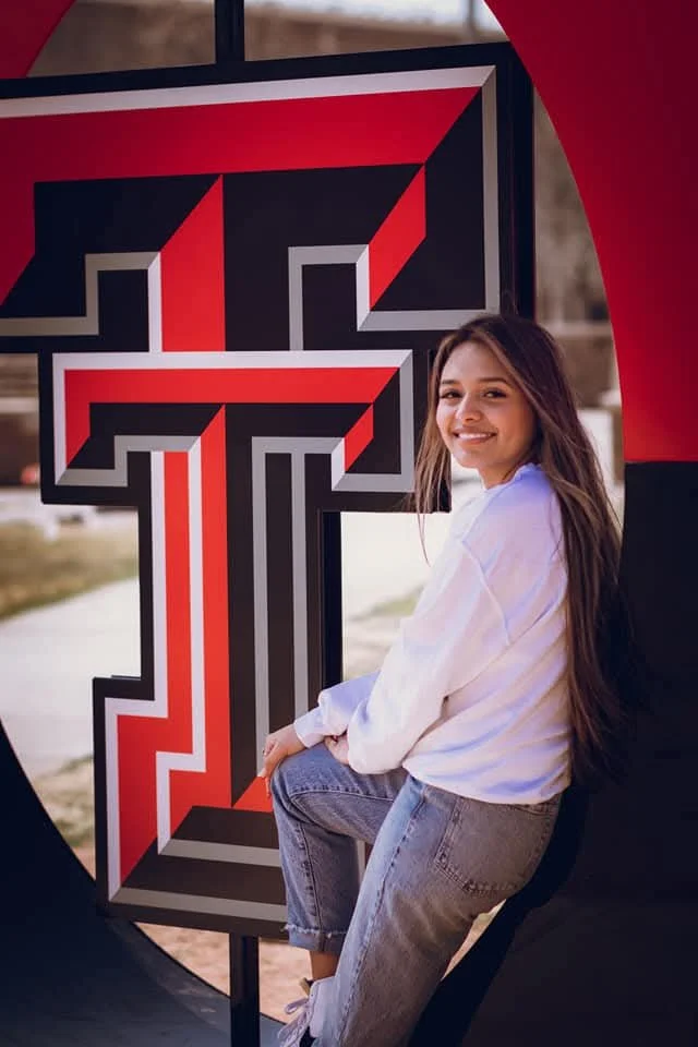 A smiling young woman in a white long sleeve shirt and jeans sitting next to a large red and black Texas Tech Double T logo sign in Lubbock Texas