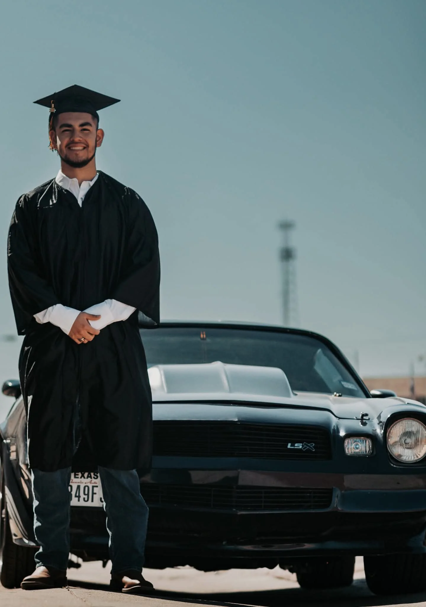 A smiling young man in a black graduation cap and gown standing next to a classic black muscle car in Lubbock Texas