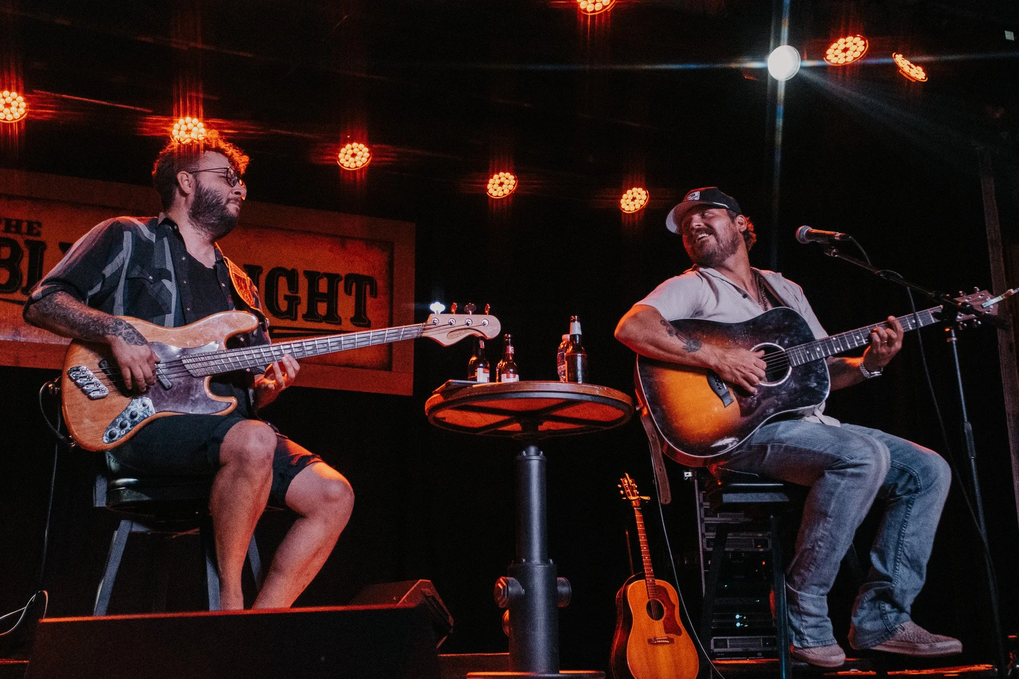 A live concert photograph of an acoustic guitarist performing on stage alongside a bass guitar player under bright warm stage lights at The Blue Light in Lubbock Texas
