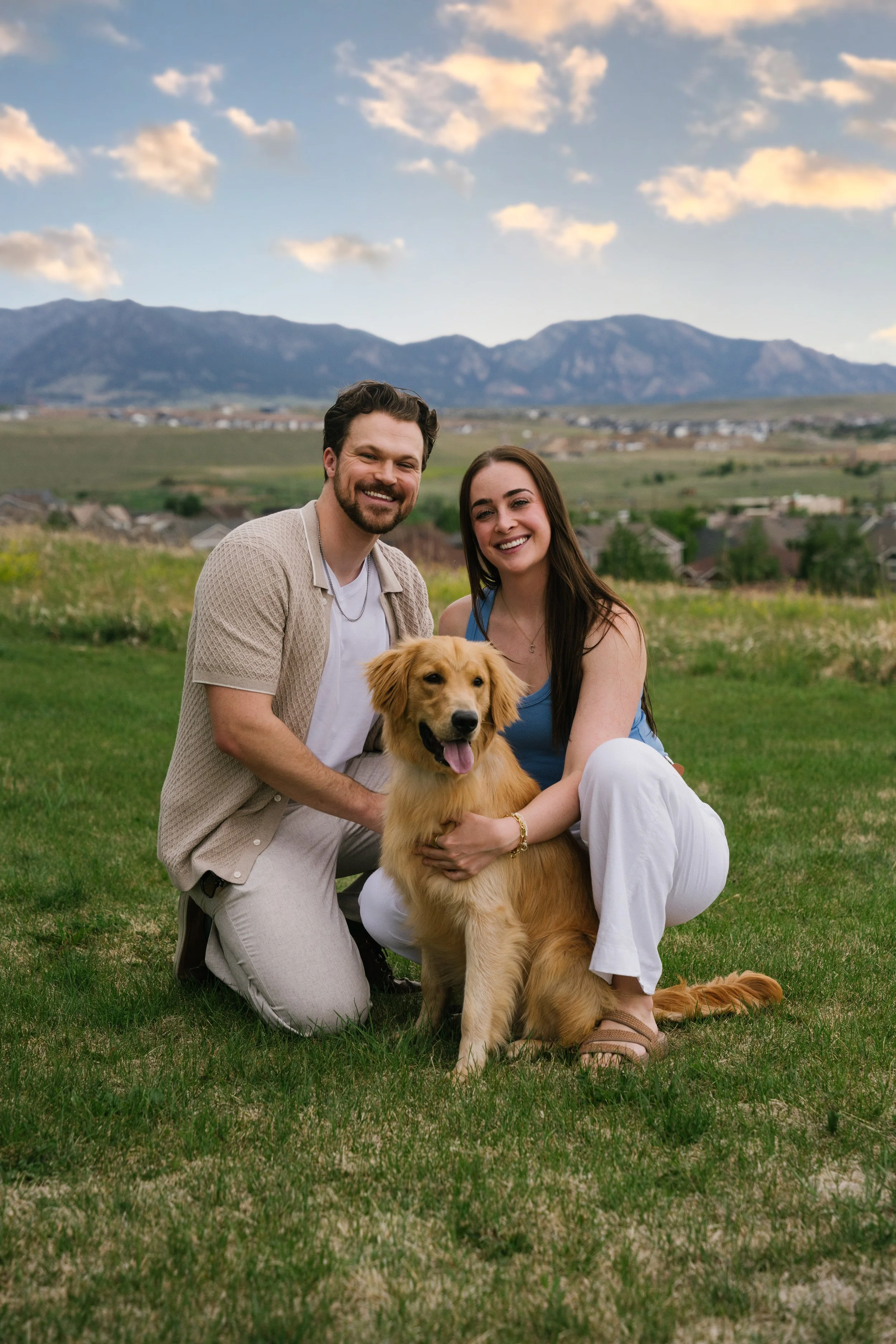A happy couple and their golden retriever dog outdoors on a grassy field with mountains and a partly cloudy sky in the background.