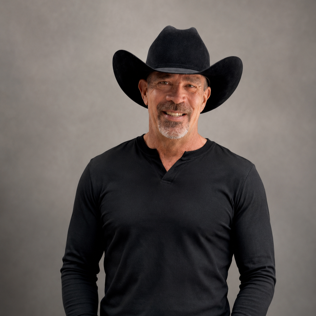 A man wearing a black cowboy hat and black long-sleeve shirt, smiling against a gray background.
