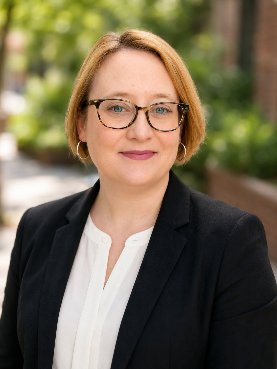 Professional headshot of a blond woman with short hair, blue eyes, wearing glasses in a white shirt and black blazer.