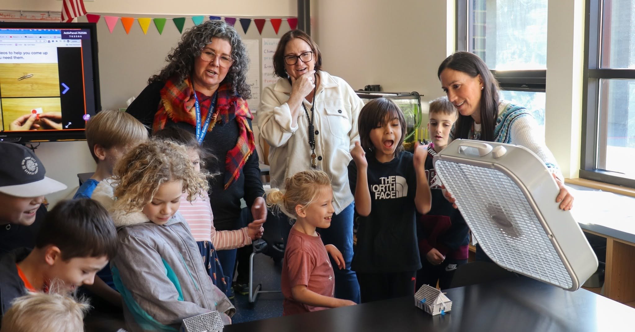 A group of students and teachers surround a table where a teacher is testing out a student's paper house. Students are excited to see the house stands while teachers smile.