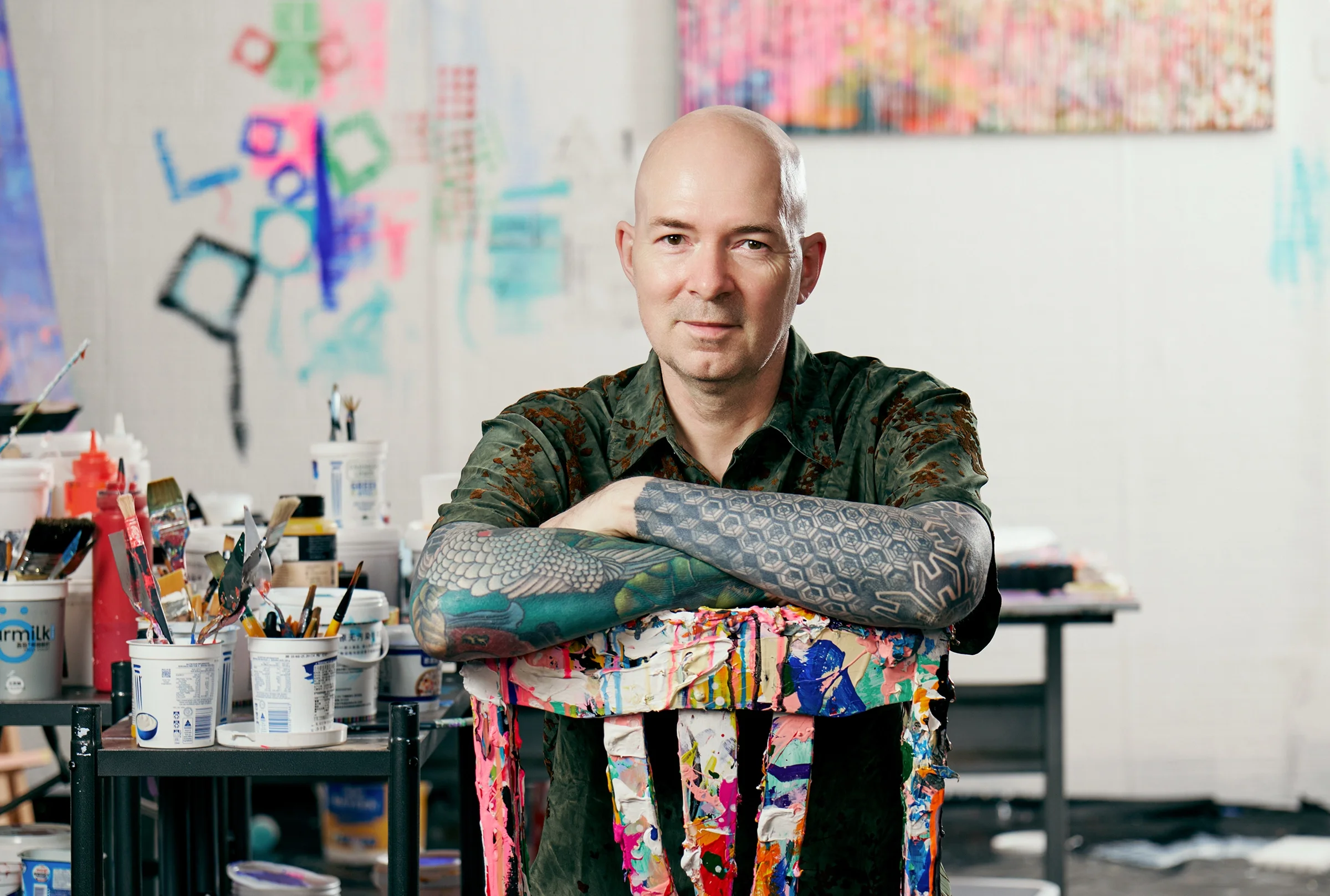 A bald man with tattoos on his arm, wearing a paint-splattered apron, sits with arms crossed in an art studio filled with various art supplies, colorful paintings, and abstract artwork on the walls.
