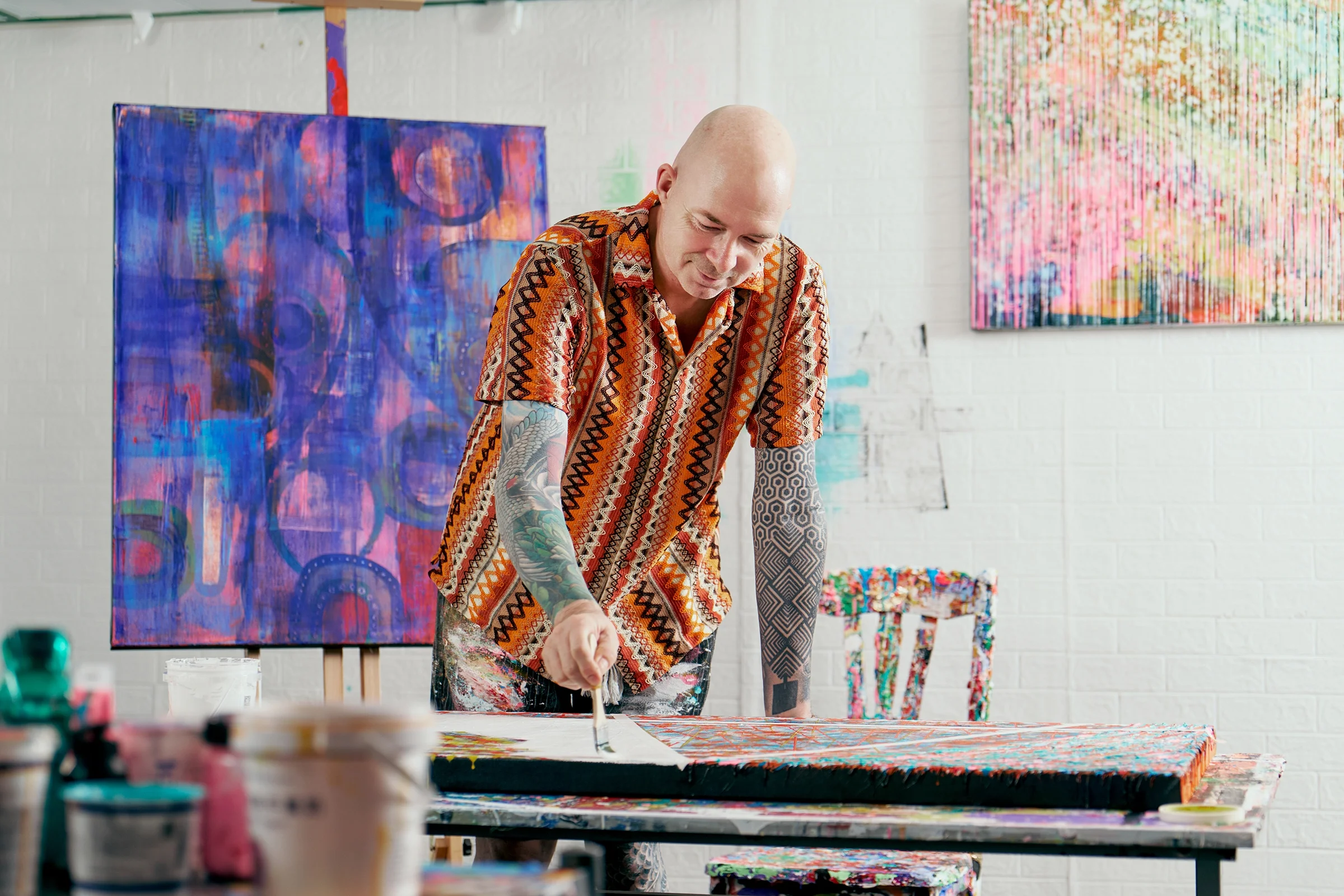 A bald man with colorful tattoos on his arms, wearing a patterned shirt, is working on a large, colorful abstract painting in an art studio with white brick walls and multiple abstract paintings in the background.