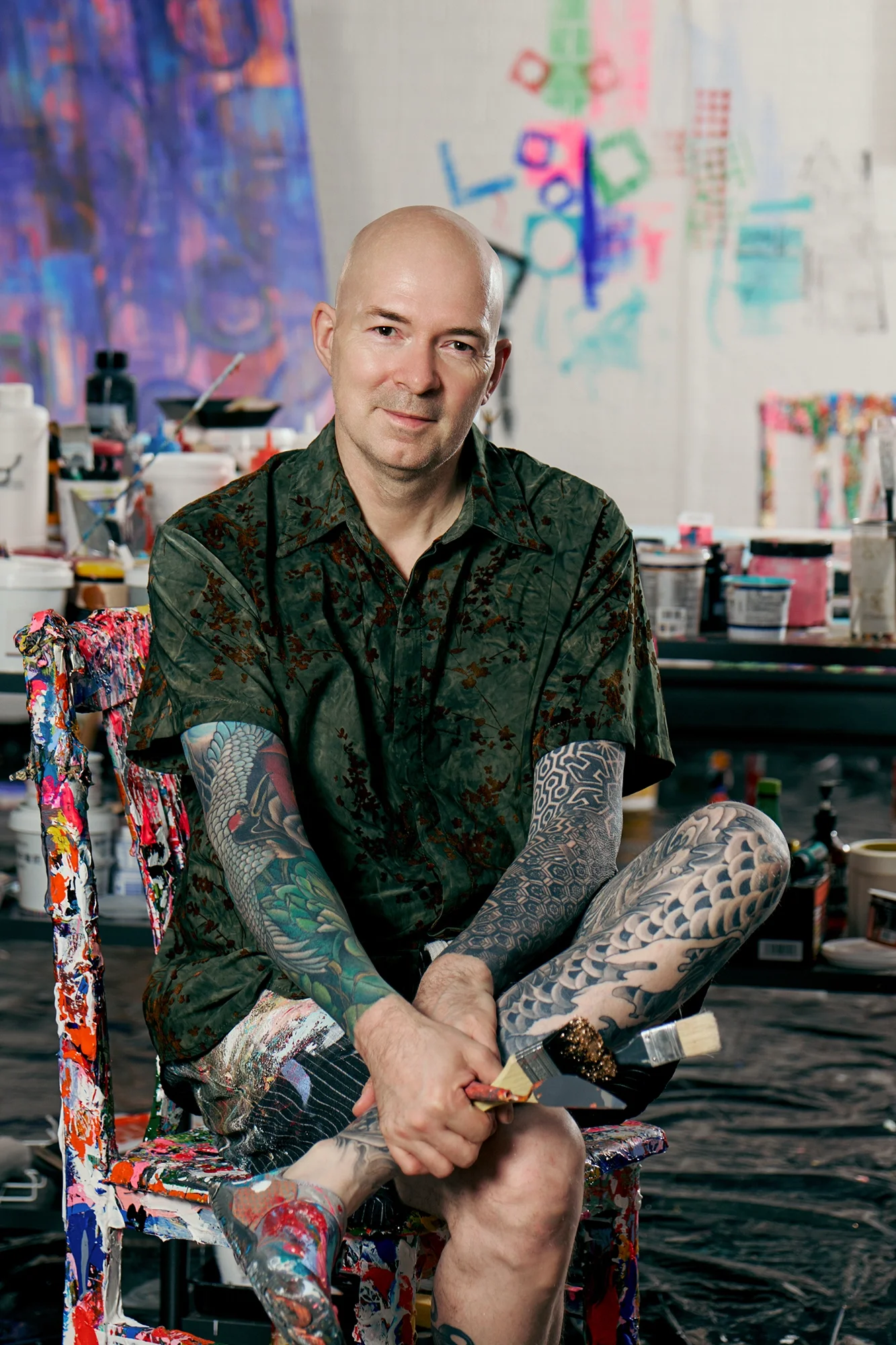 An artist with tattoos sitting on a colorful paint-splattered chair in a studio surrounded by paints and canvases, holding a paintbrush.