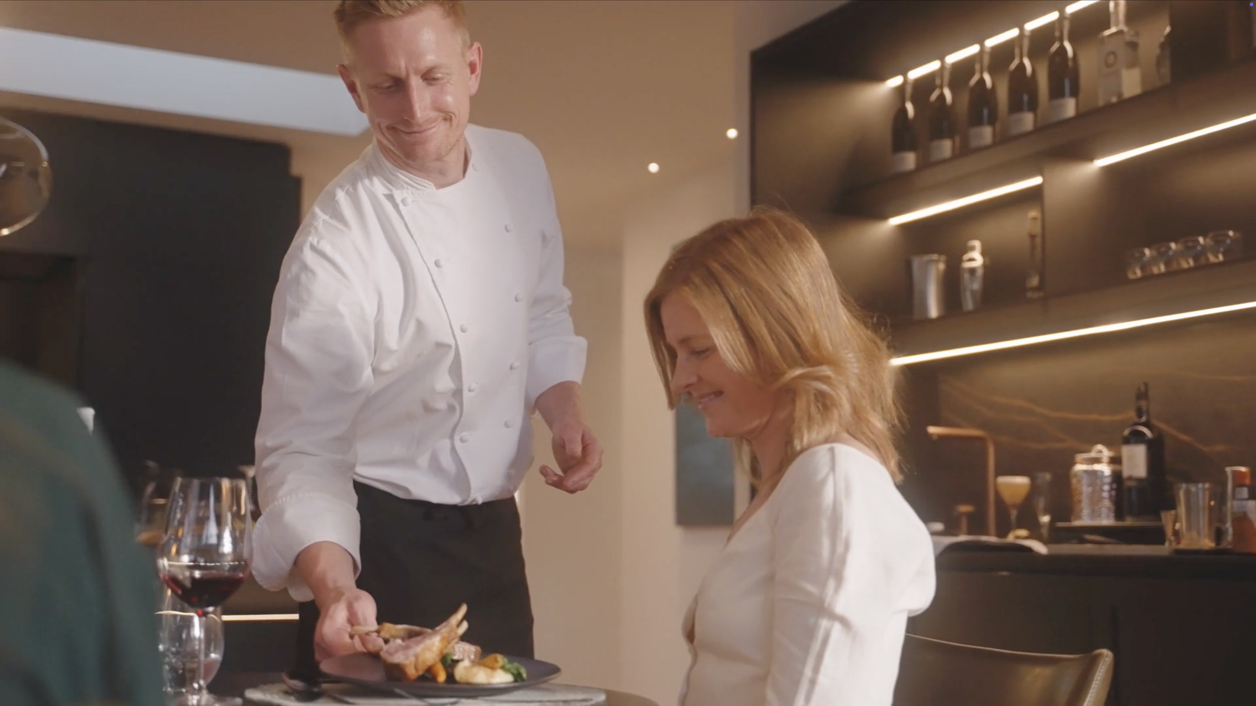 A chef in a white uniform serving a woman at a restaurant. The woman, with reddish hair, is smiling, and there is a wine glass in front of her. The background shows wine bottles and kitchen shelves.