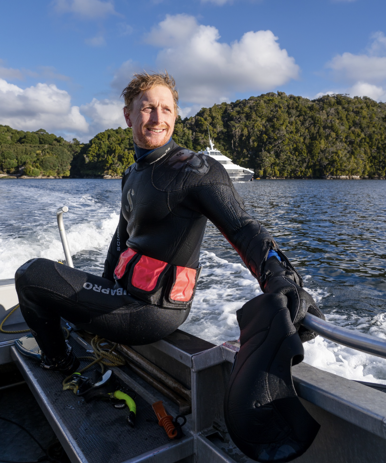 Smiling man in a wetsuit on a boat, with a yacht and green islands in the background, enjoying a sunny day on the water.