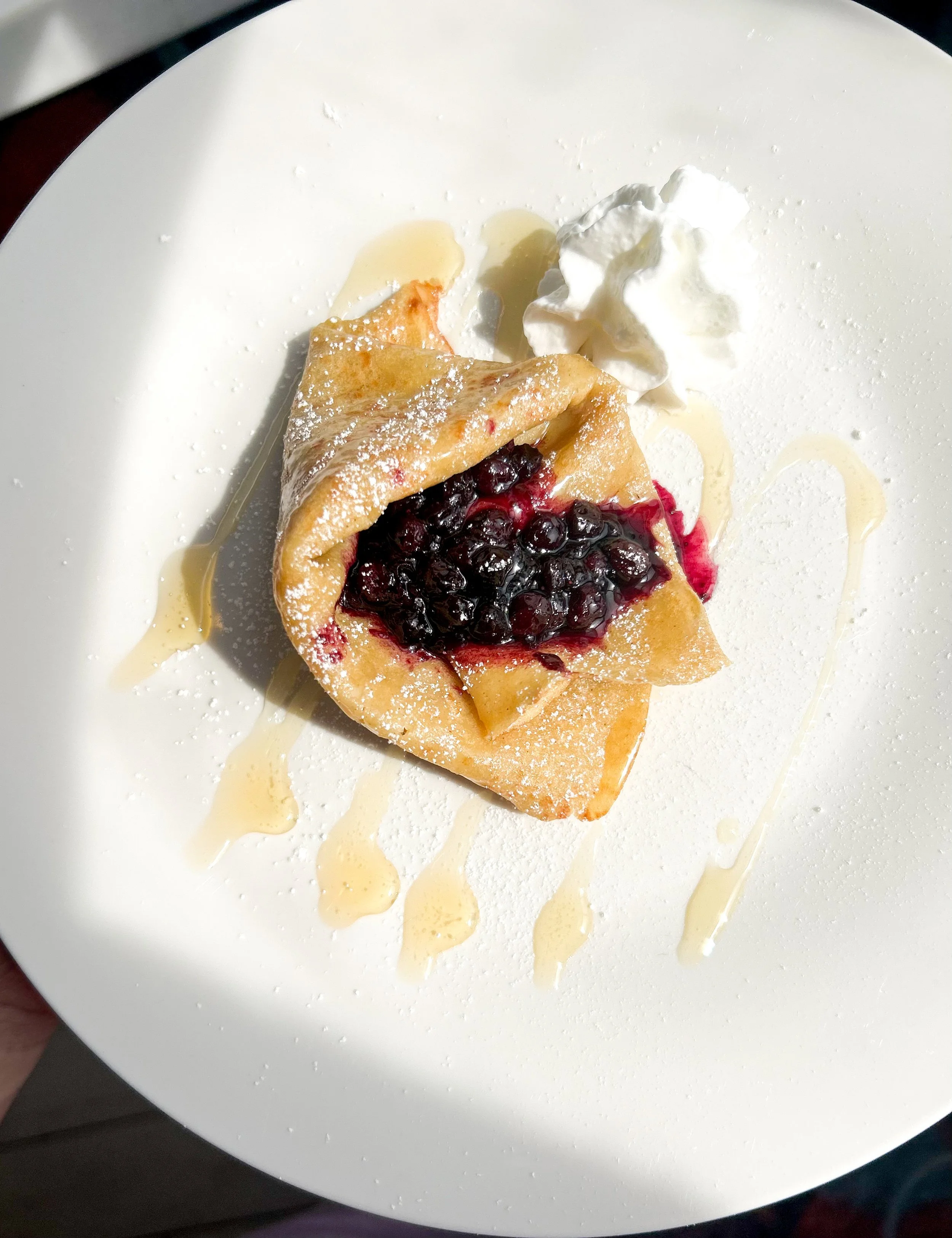 A dessert of a slice of flaky pie filled with blueberries, topped with whipped cream, drizzled with honey, and dusted with powdered sugar on a white plate.