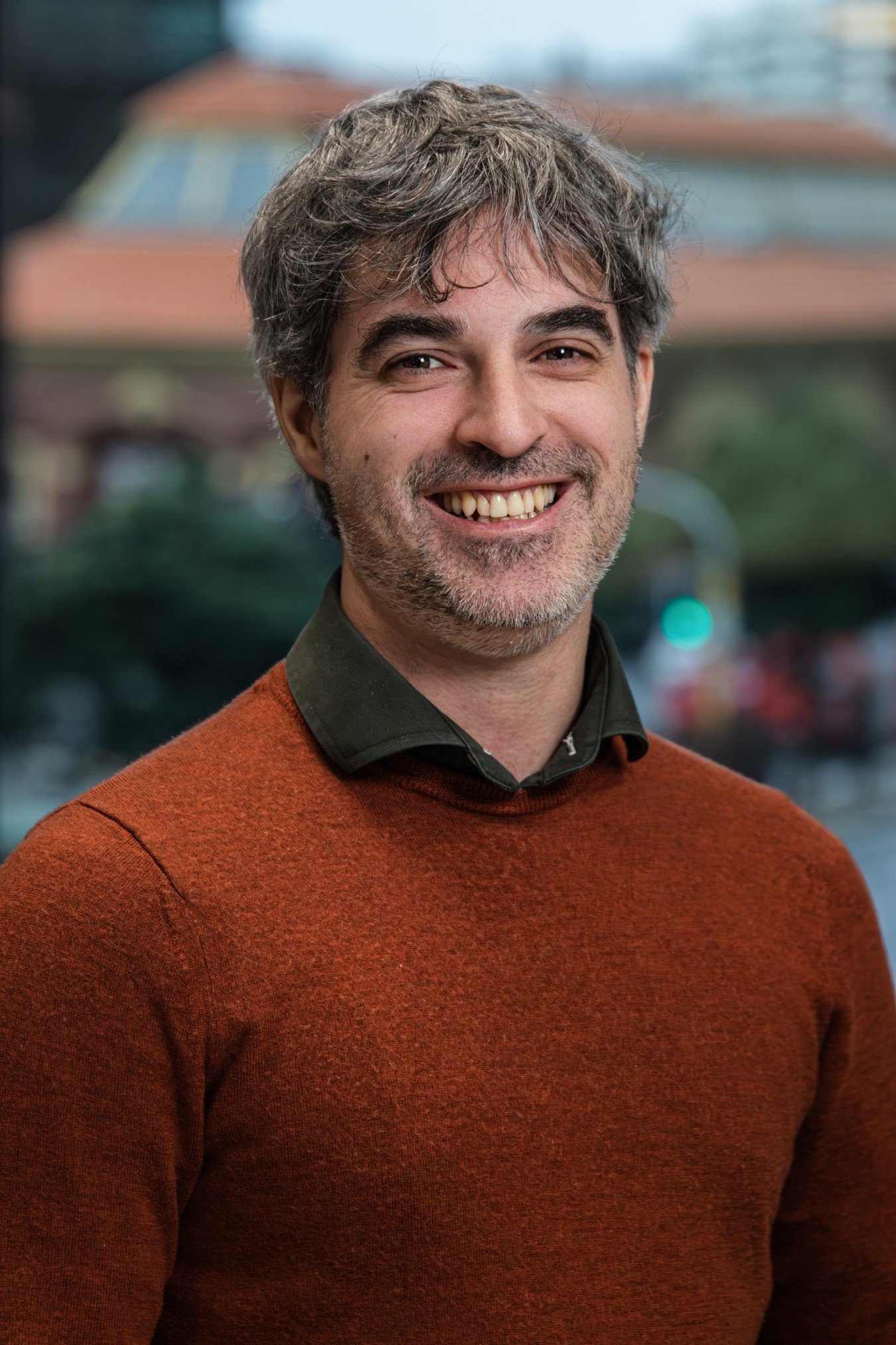 A man with brown hair and a beard smiling, wearing an orange sweater and a dark shirt, with a blurred cityscape background.