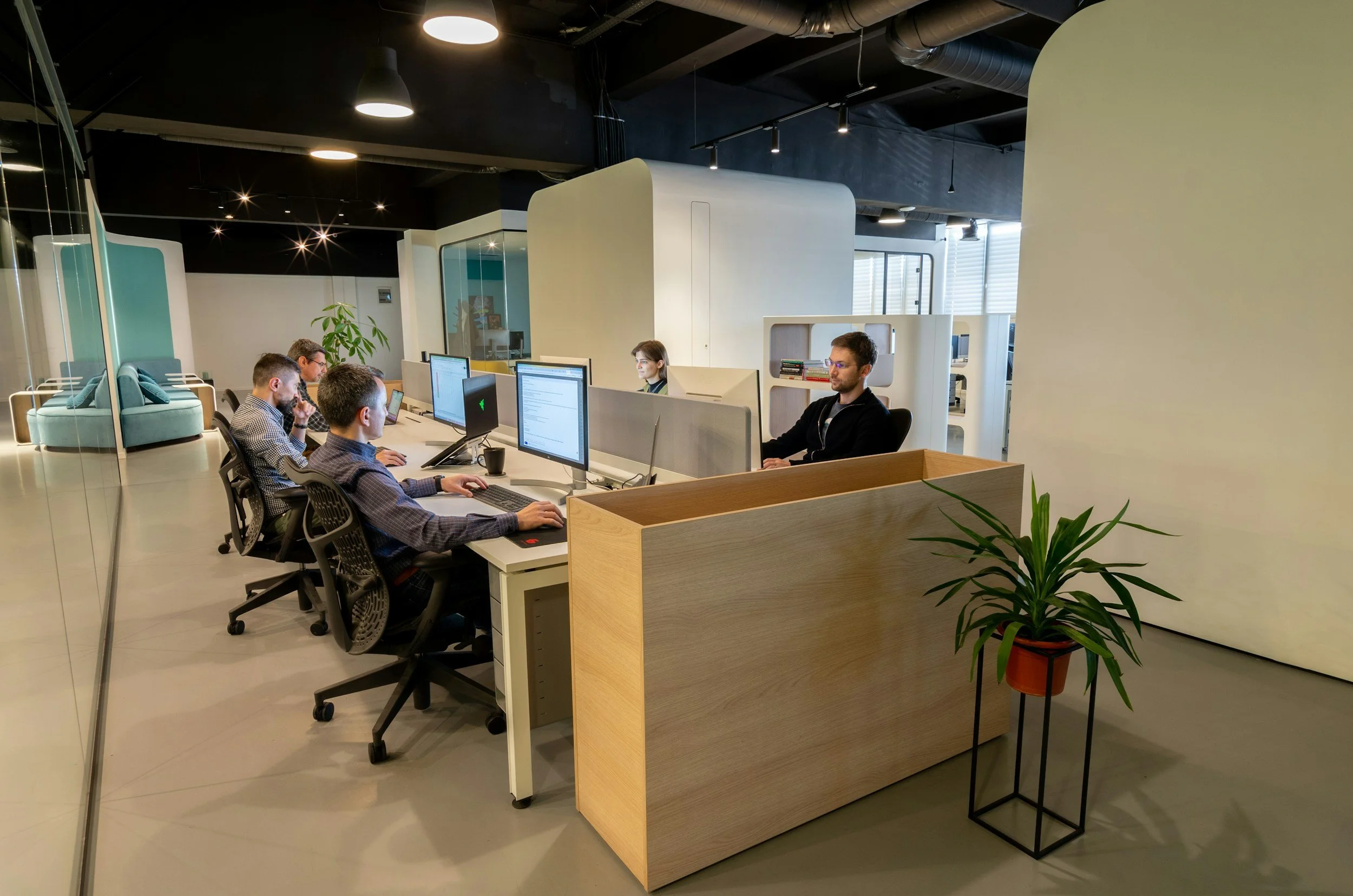 Office workspace with multiple employees working at desks with desktop computers. The office has modern, open design with glass walls, indoor plants, and ample natural light.