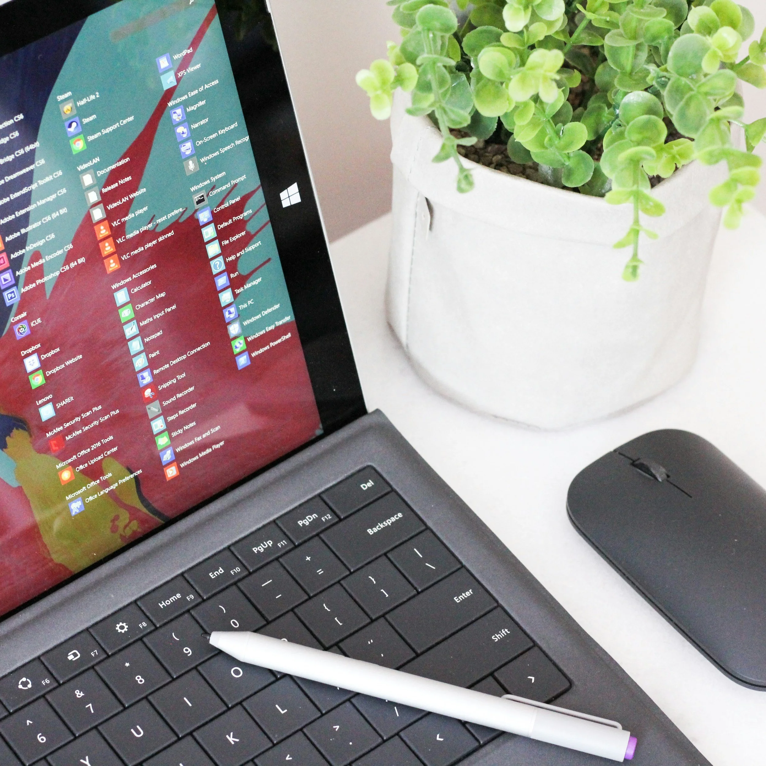 Close-up of a black laptop keyboard with a white stylus resting on it, and part of the laptop screen showing a colorful desktop with various icons. A white flowerpot with a green succulent plant is positioned next to the laptop.