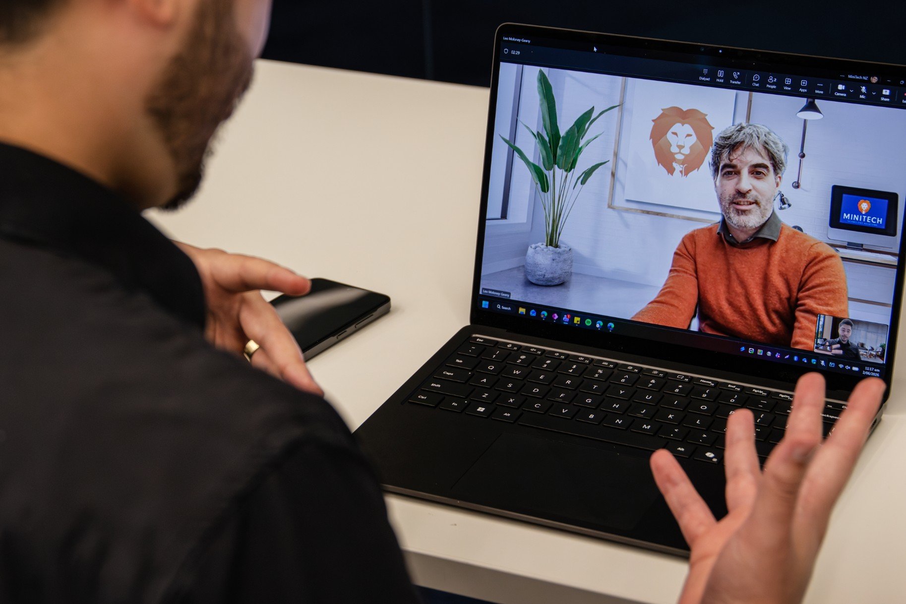 Person participating in a video call on a laptop, showing a man with gray hair and beard in a cozy home office with artwork and a plant in the background.