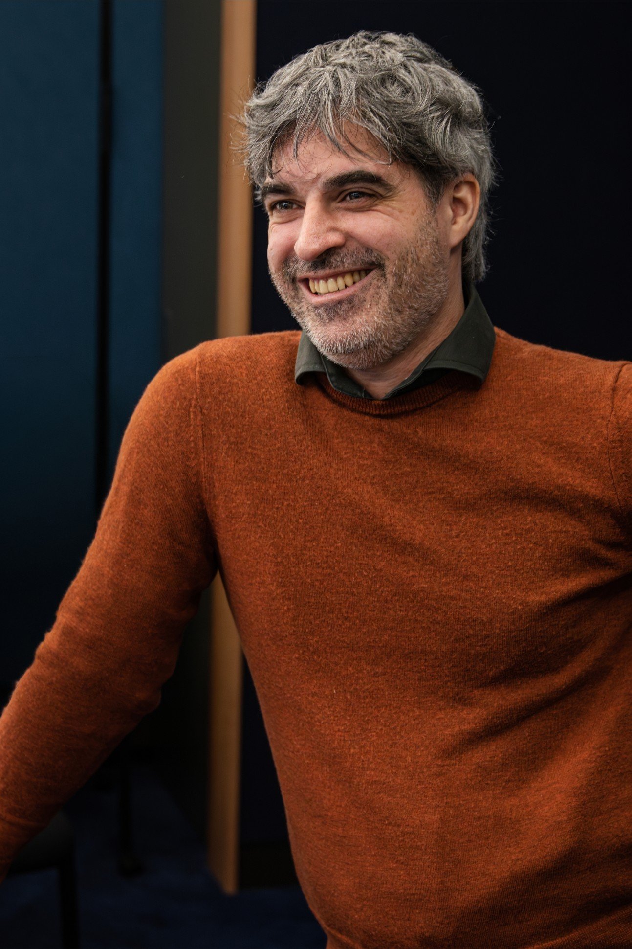 Close-up of a smiling man with gray hair and beard, wearing an orange sweater over a dark collared shirt, sitting indoors.