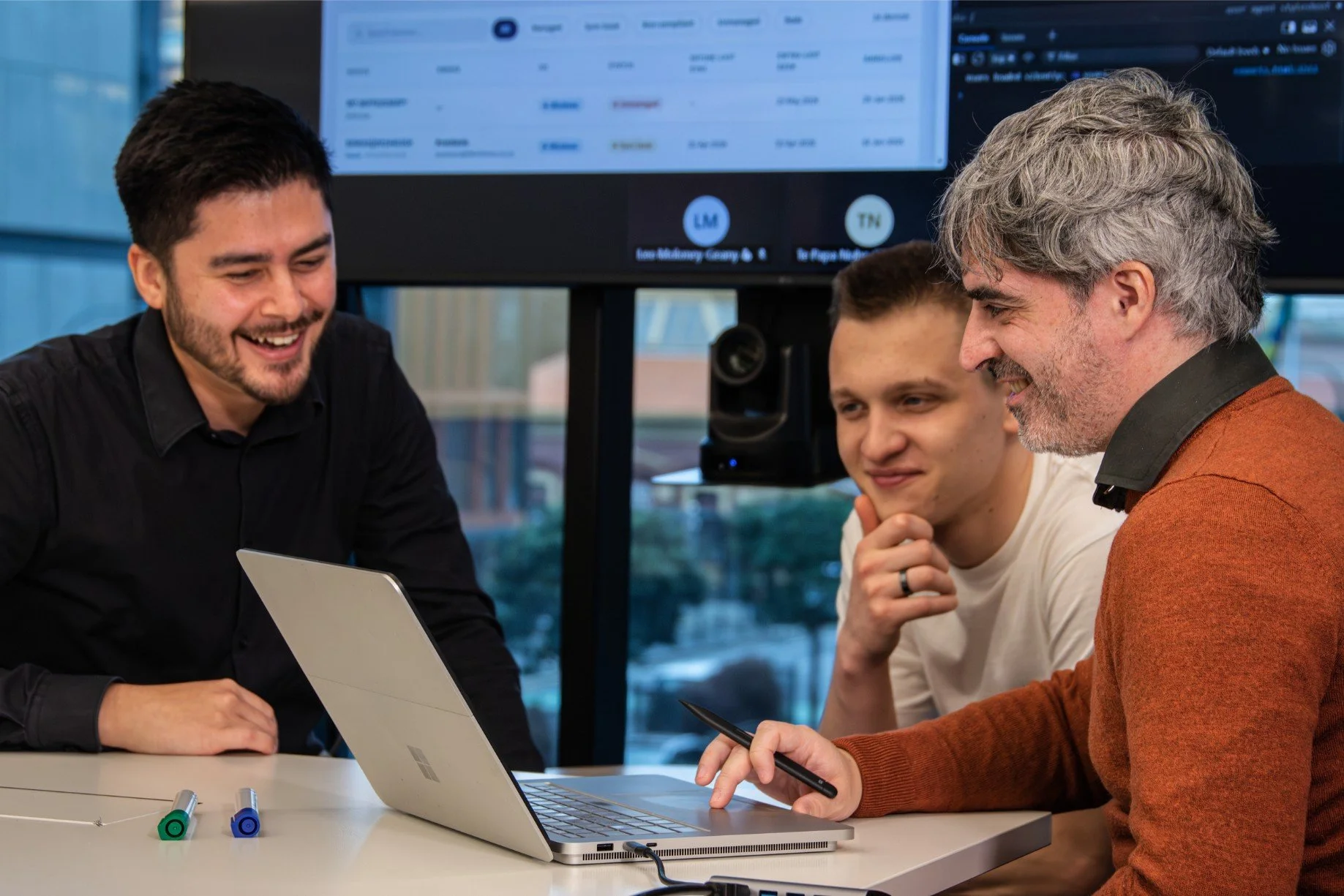 Three men are sitting at a table with a laptop, engaged in a discussion, in a modern office setting.