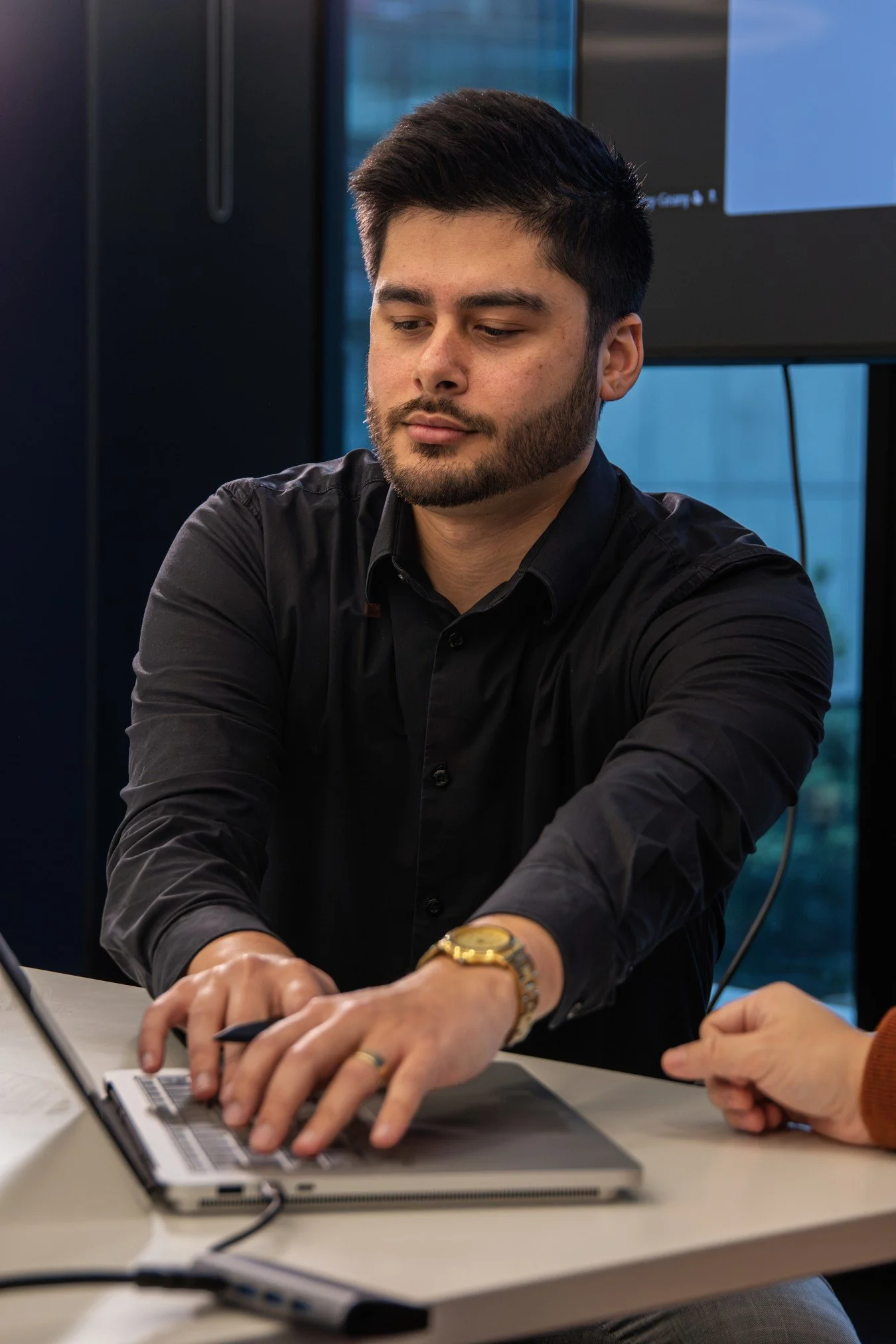 A man with dark hair and a beard, wearing a black shirt and a gold watch, working on a laptop at a table in a modern office environment.